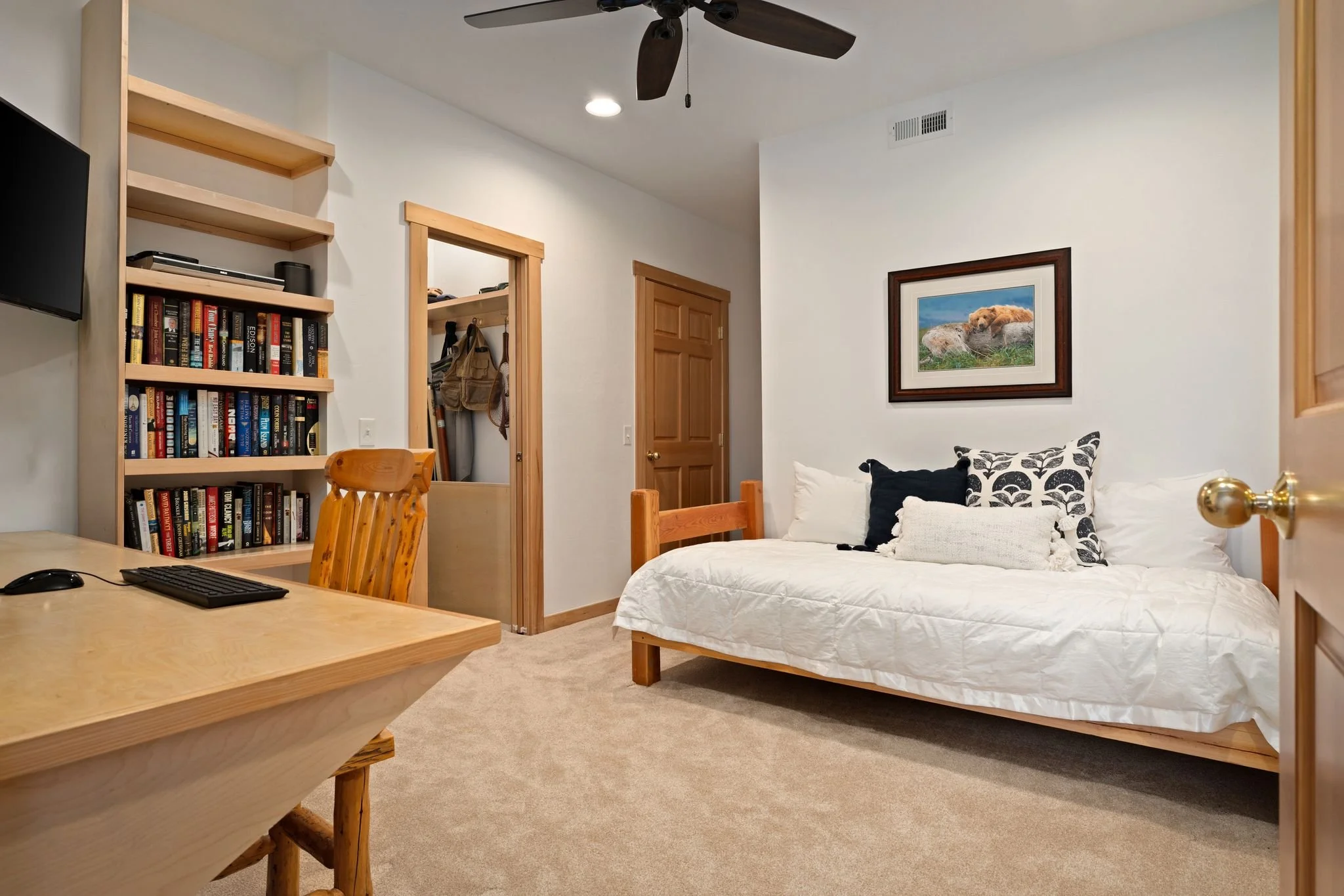 A cozy bedroom with a daybed covered in white bedding, decorative pillows, a framed nature photograph on the wall, a bookshelf filled with books, a wooden desk with a keyboard and mouse, a ceiling fan, and an open closet door showing hanging clothes.