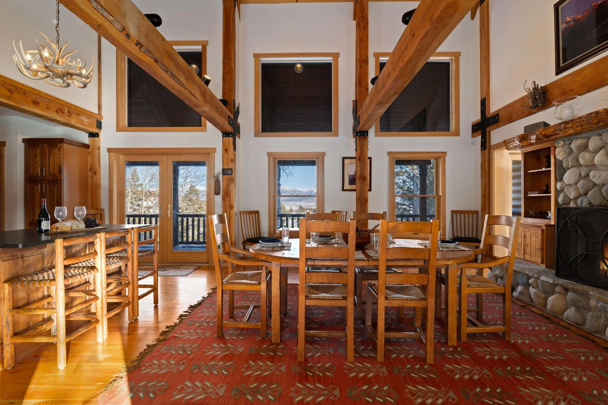 A rustic dining room with high ceilings, wooden beams, and large windows showing trees and mountains outside. It features a round wooden dining table with eight matching chairs, a stone fireplace with a fire burning, and a red patterned area rug. The