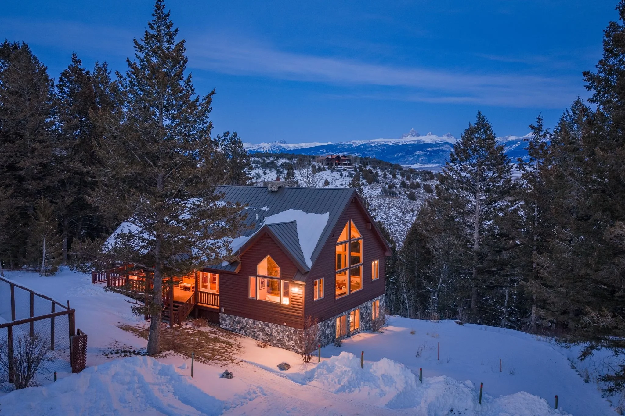 A snow-covered house with warm interior lighting surrounded by trees in a mountainous landscape during dusk.