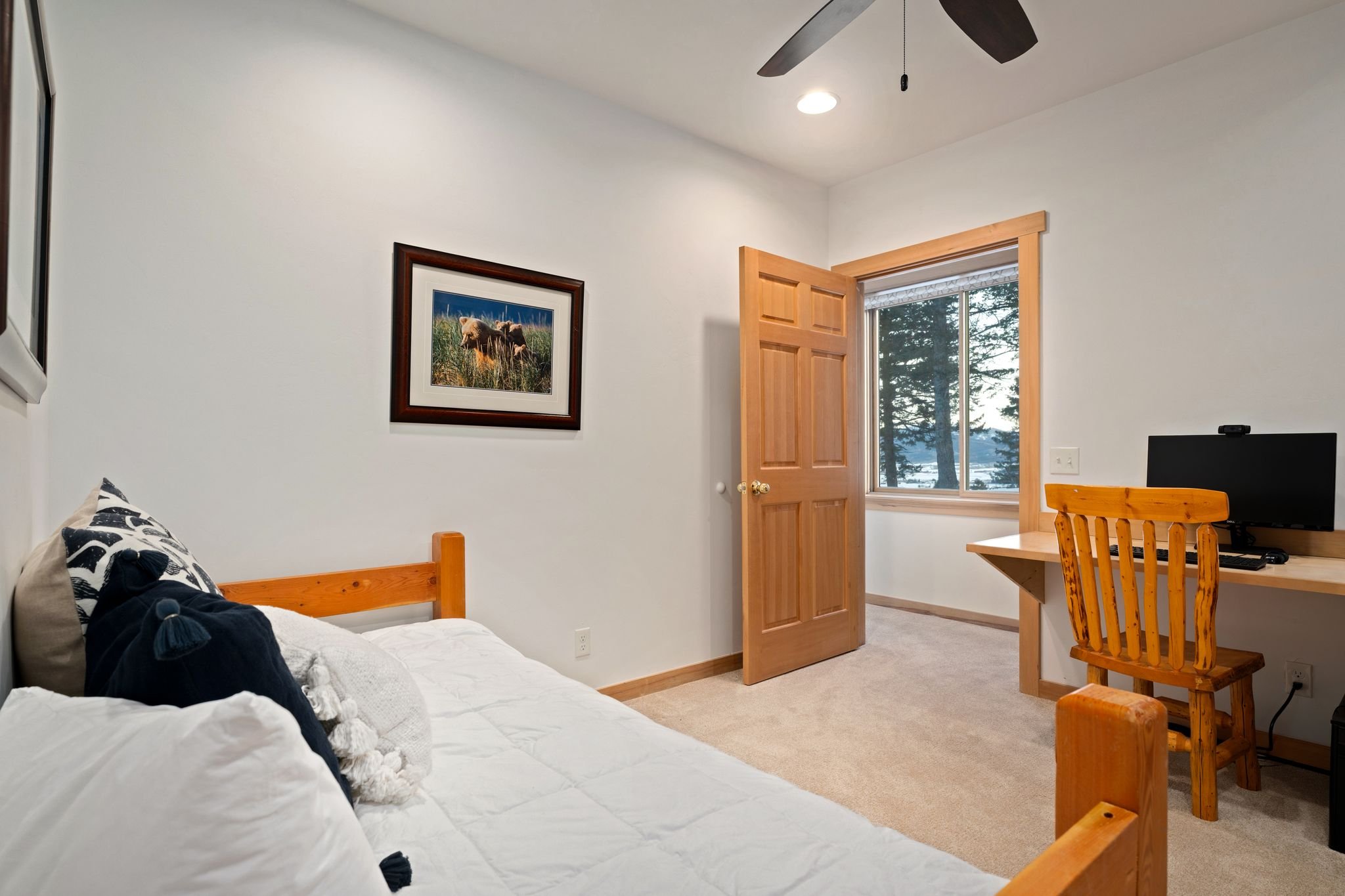 A bedroom with a bed on the left, a desk with a computer and a chair on the right, a window showing trees outside, a ceiling fan, a painting on the wall, and a wooden door.