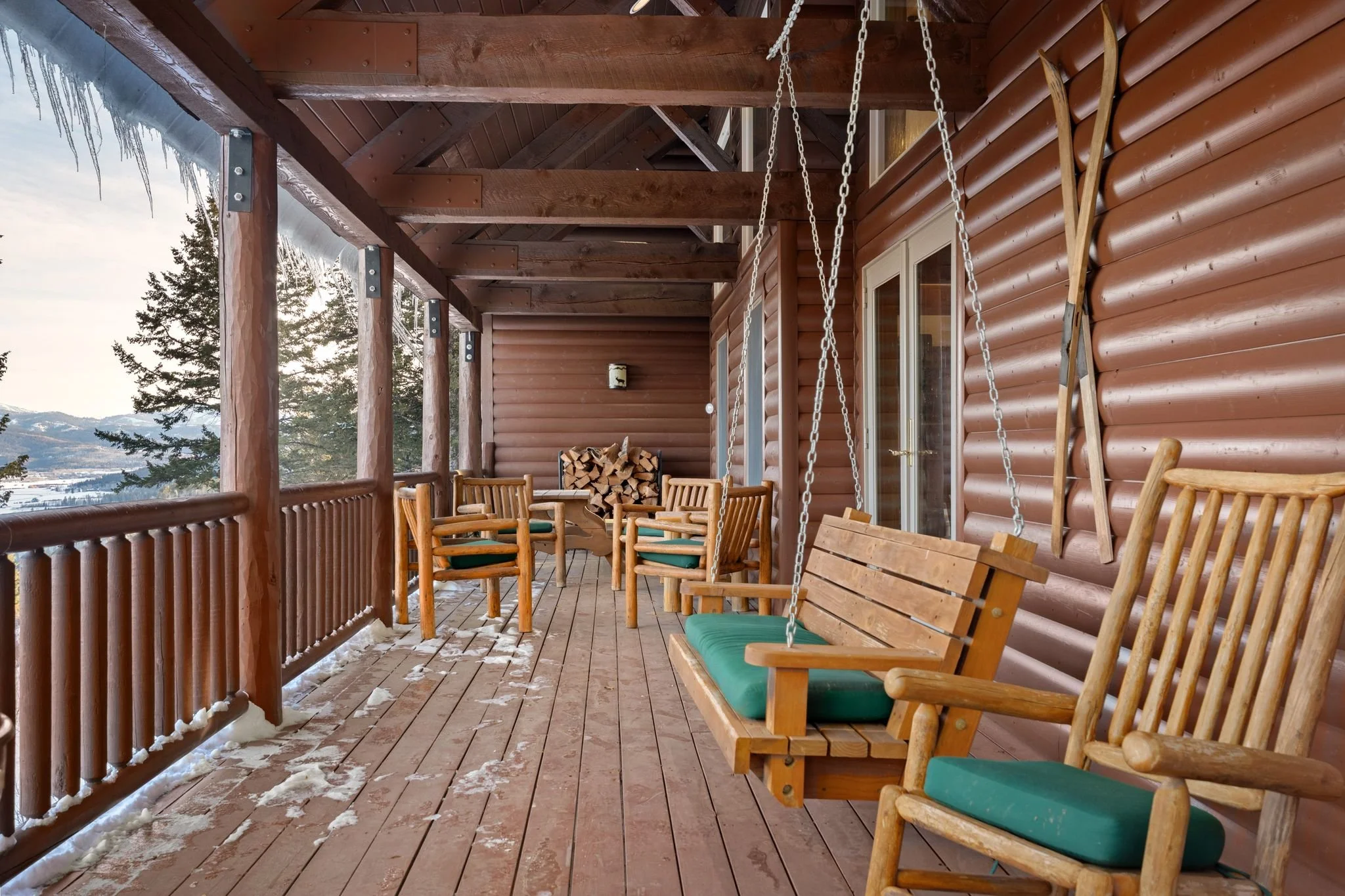 A covered wooden porch with wooden chairs, a swing, and a stack of firewood. Snow is on the ground, and snow-covered mountains and trees are visible in the background.