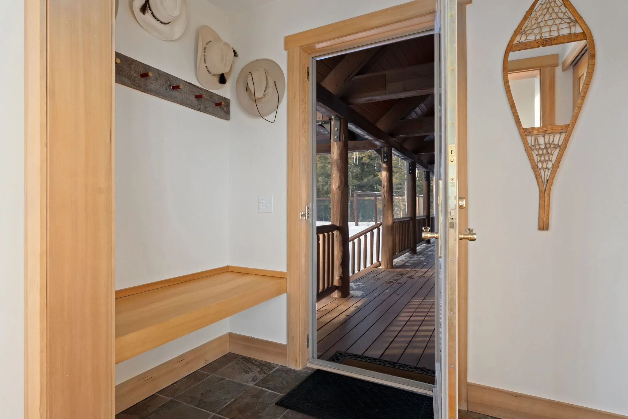 Interior entryway with a wooden bench, beige hats on a wall-mounted coat rack, an oval mirror, and a glass door leading to a covered wooden porch with wooden support beams.