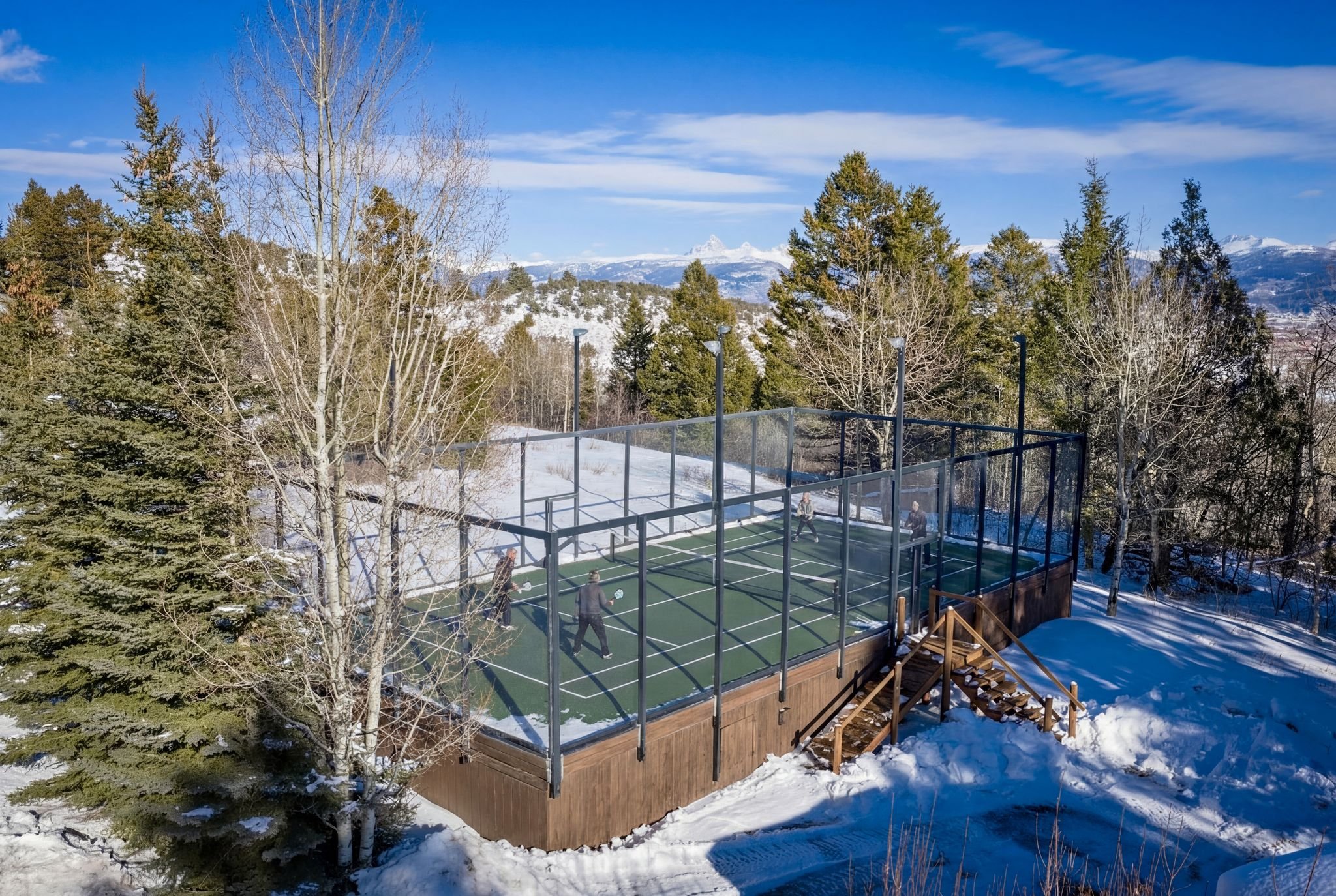 Outdoor heated pickleball court with players, surrounded by snow-covered ground and trees, mountainous landscape in the background.