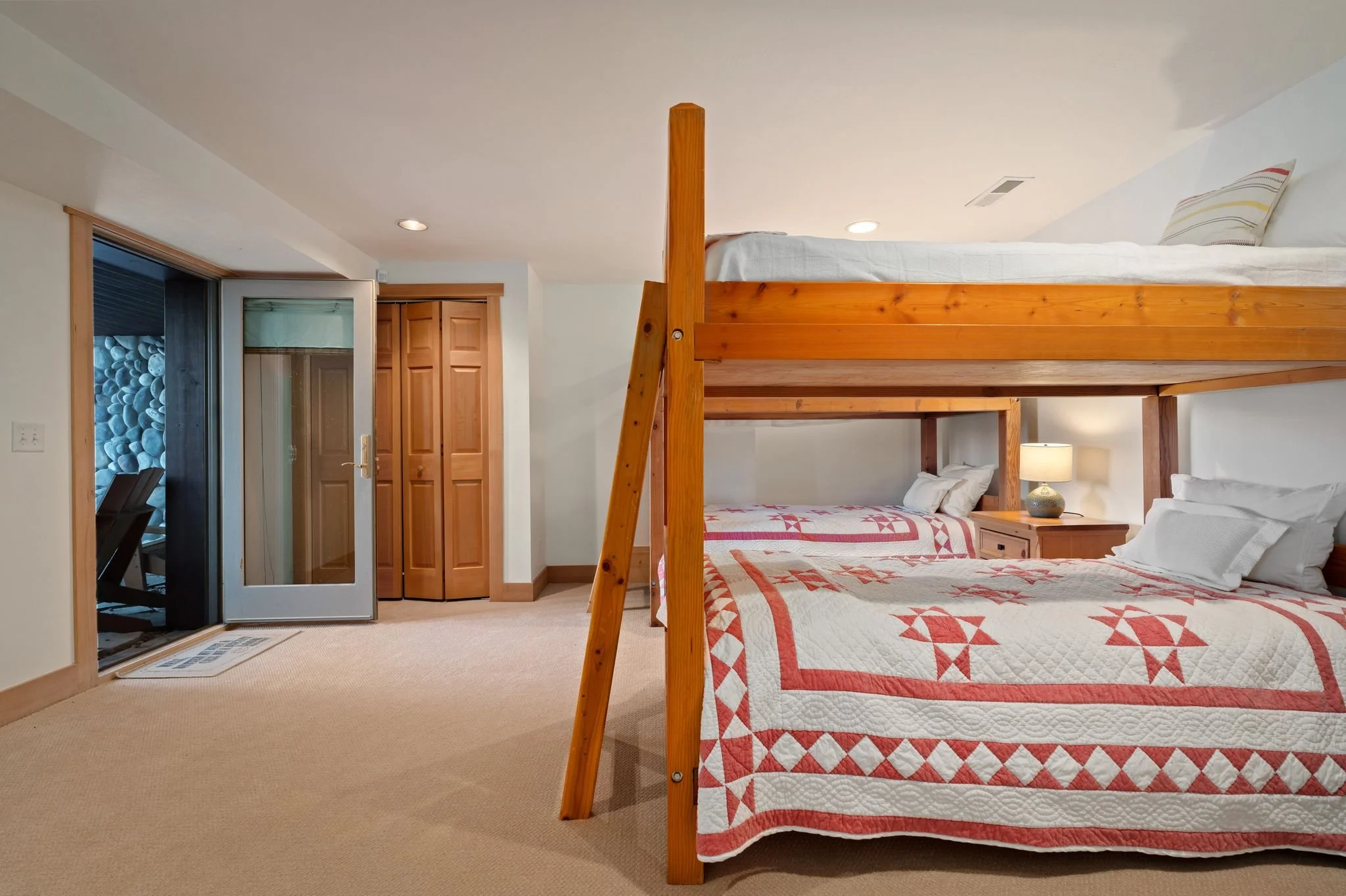 A bedroom with white walls, beige carpet, and a wooden bunk bed. The lower bed has a quilt with a red and white geometric pattern. There is a nightstand with a lamp next to it. A door with a glass panel leads to an outdoor area, and there are closet 