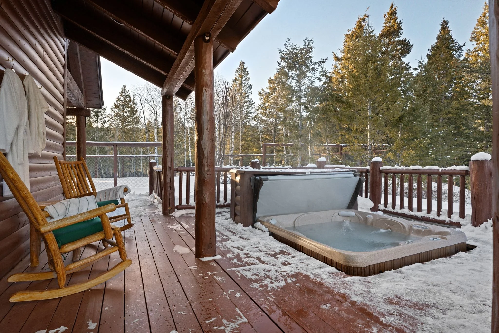A snowy outdoor deck of a log cabin with wooden rocking chairs, a hot tub, and a snowy landscape with tall pine trees in the background.
