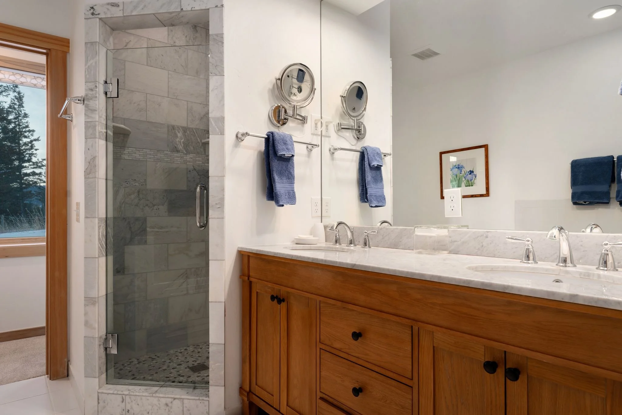 Bathroom showing a glass-enclosed shower, wooden vanity with marble countertop, and a large mirror. Blue towels hang on the wall, and a small framed picture is above the towels. A window with a view of trees is also visible.