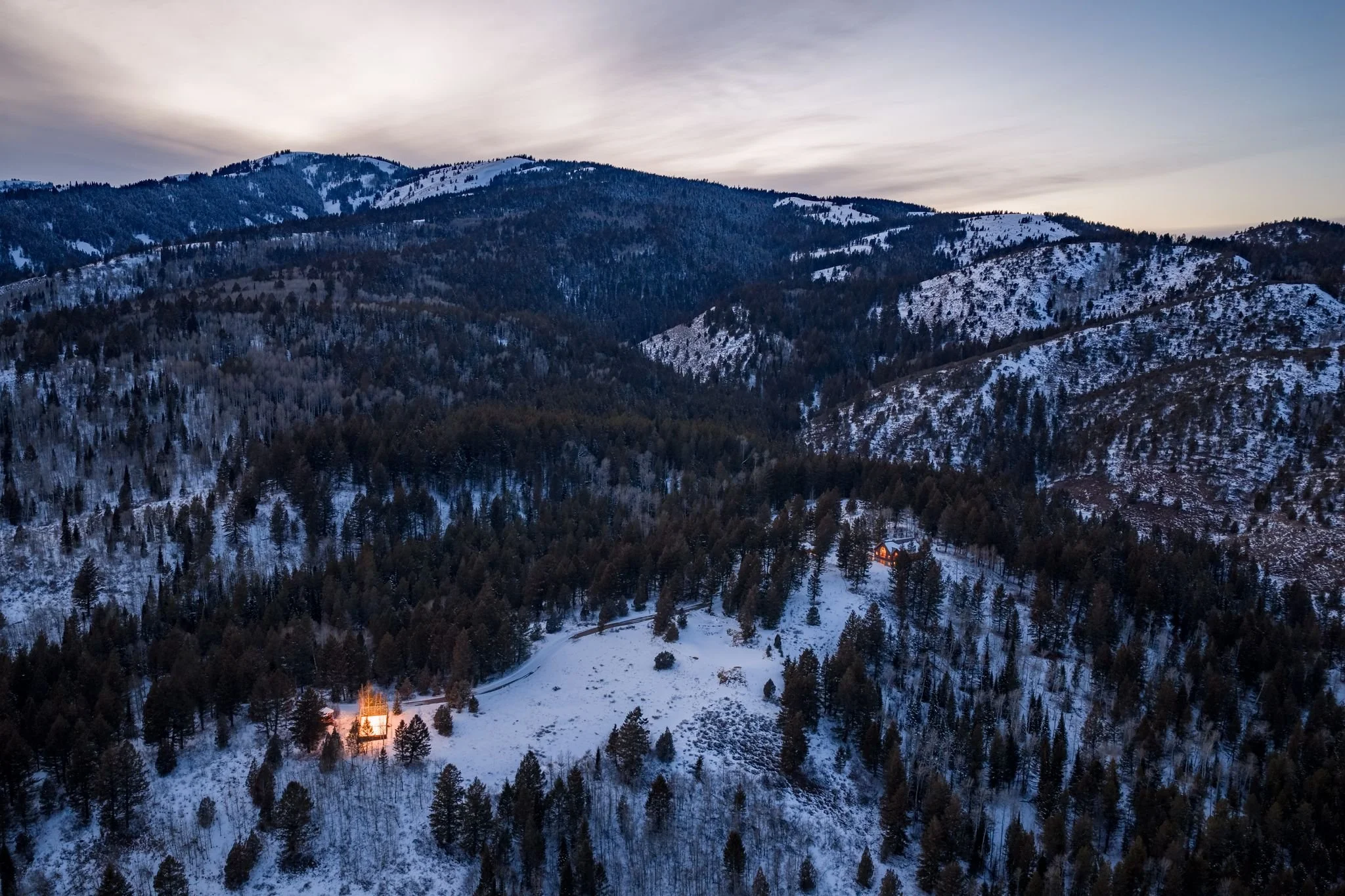 Aerial view of snow-covered mountains with pine trees during dusk, with two illuminated houses nestled among the trees.