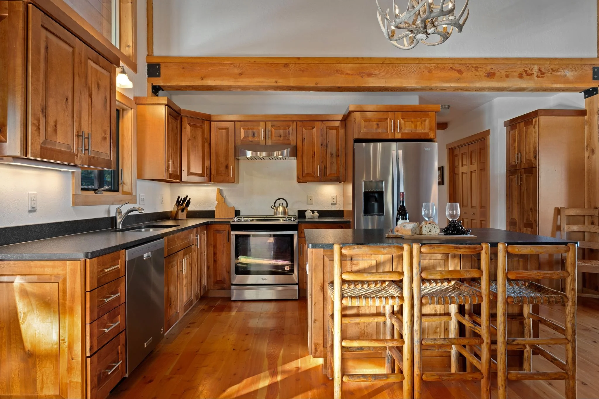 A rustic kitchen with wooden cabinets, a black countertop, stainless steel appliances, a kitchen island with bar stools, and wooden beams.