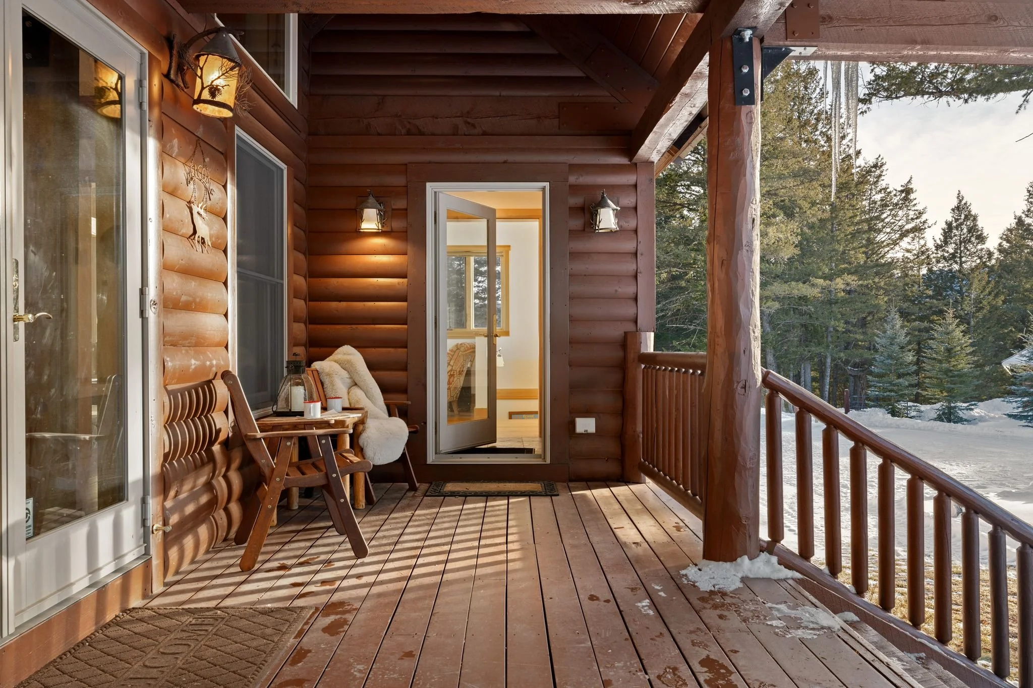 Wooden cabin porch with snow on floor, outdoor view of trees and snow, open door showing a cozy interior, outdoor seating with pillows, wall lanterns, and railing.