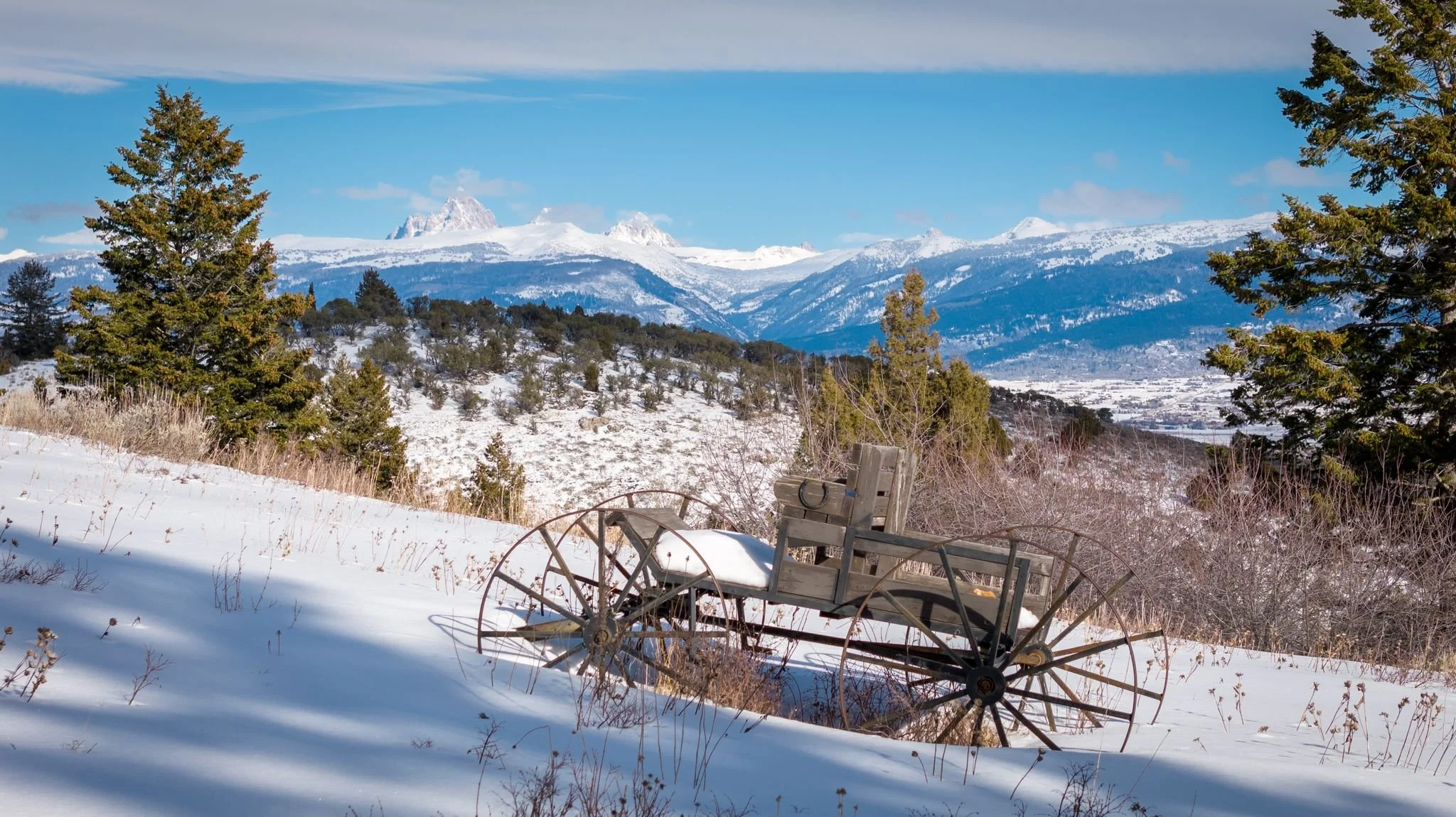 Snow-covered landscape with a rustic wooden wagon in the foreground, surrounded by evergreen trees, with snow-capped mountains and a blue sky in the background.