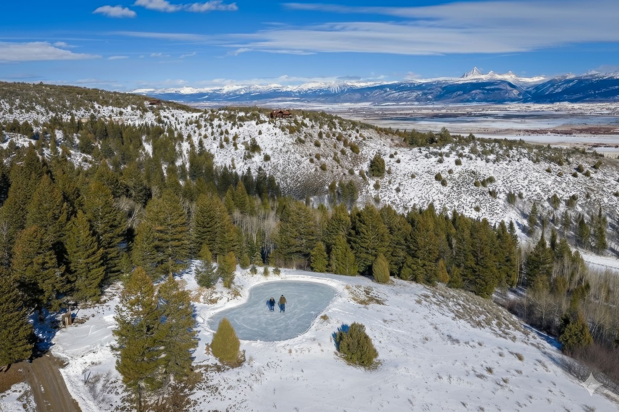 Two people ice skating on a frozen pond surrounded by snow-covered ground and green pine trees in a mountainous landscape with snow-capped peaks in the distance.
