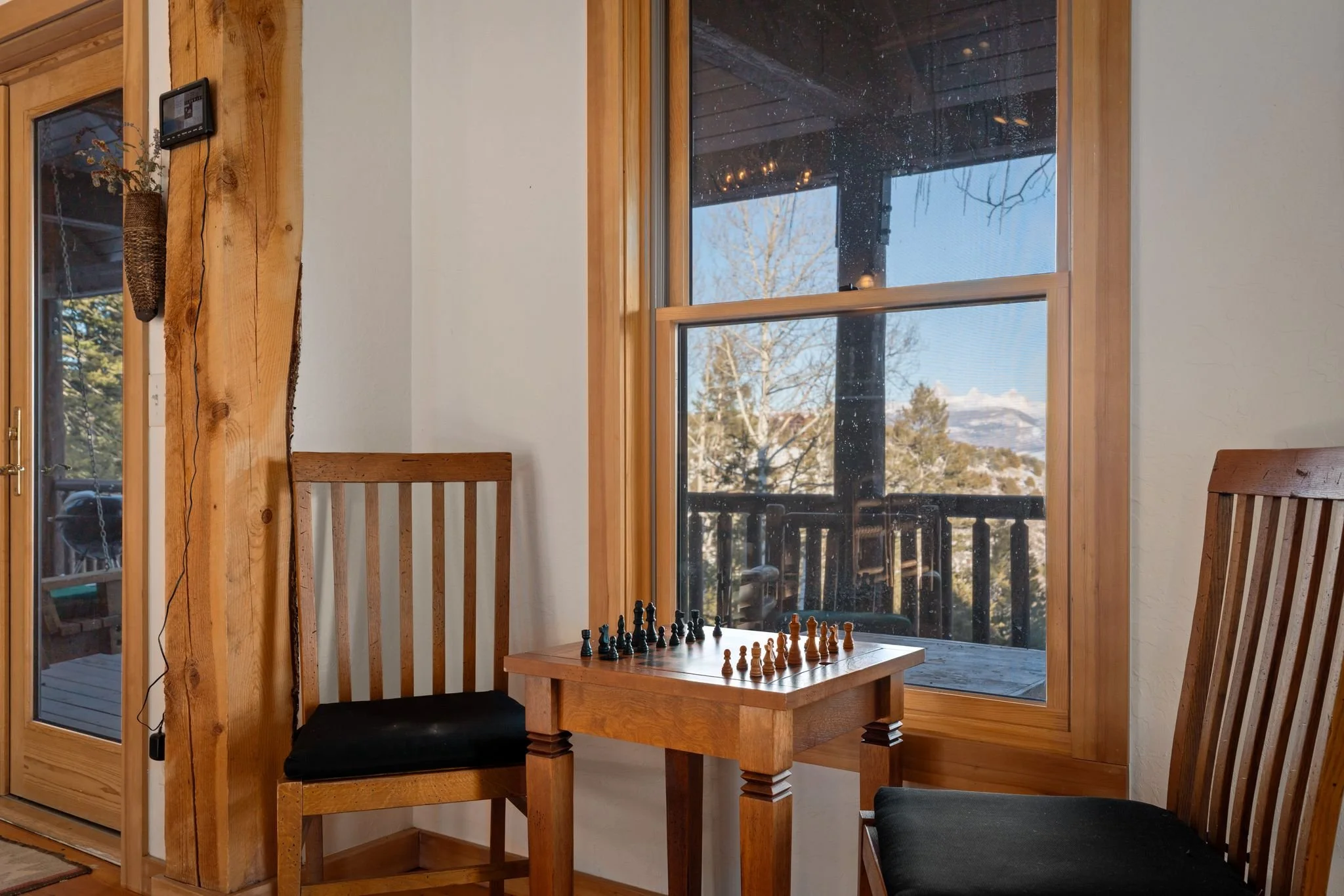 A cozy corner of a room with wooden furniture, including two chairs with black cushions and a small wooden table with a chessboard set up, next to a large window with a view of trees and a deck outside.