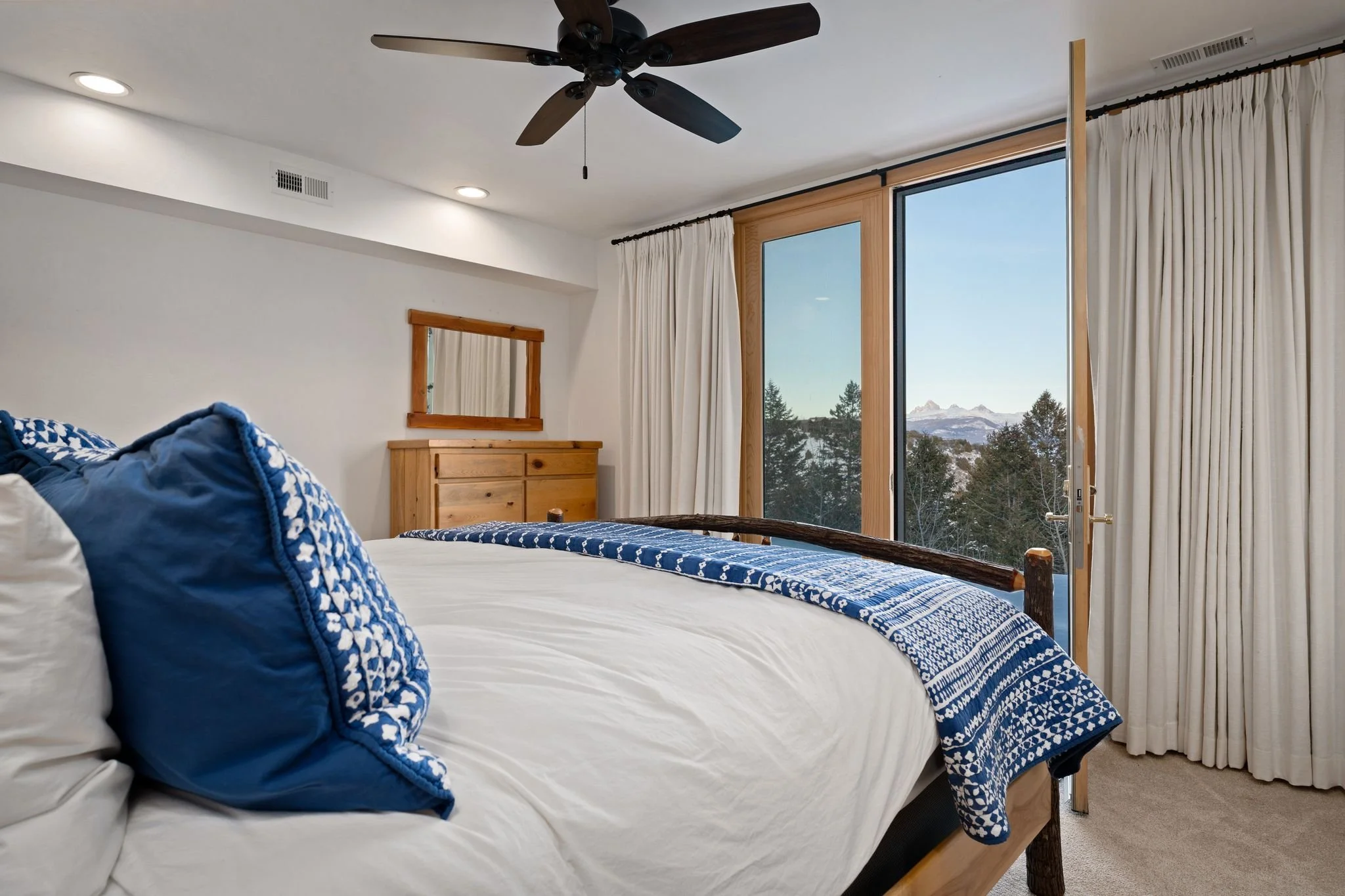 A cozy bedroom with a wooden bed frame, white bedding, and blue patterned pillows. Large sliding glass door shows mountain and forest view with snow-capped peaks in the distance, white curtains, ceiling fan, and recessed lighting.