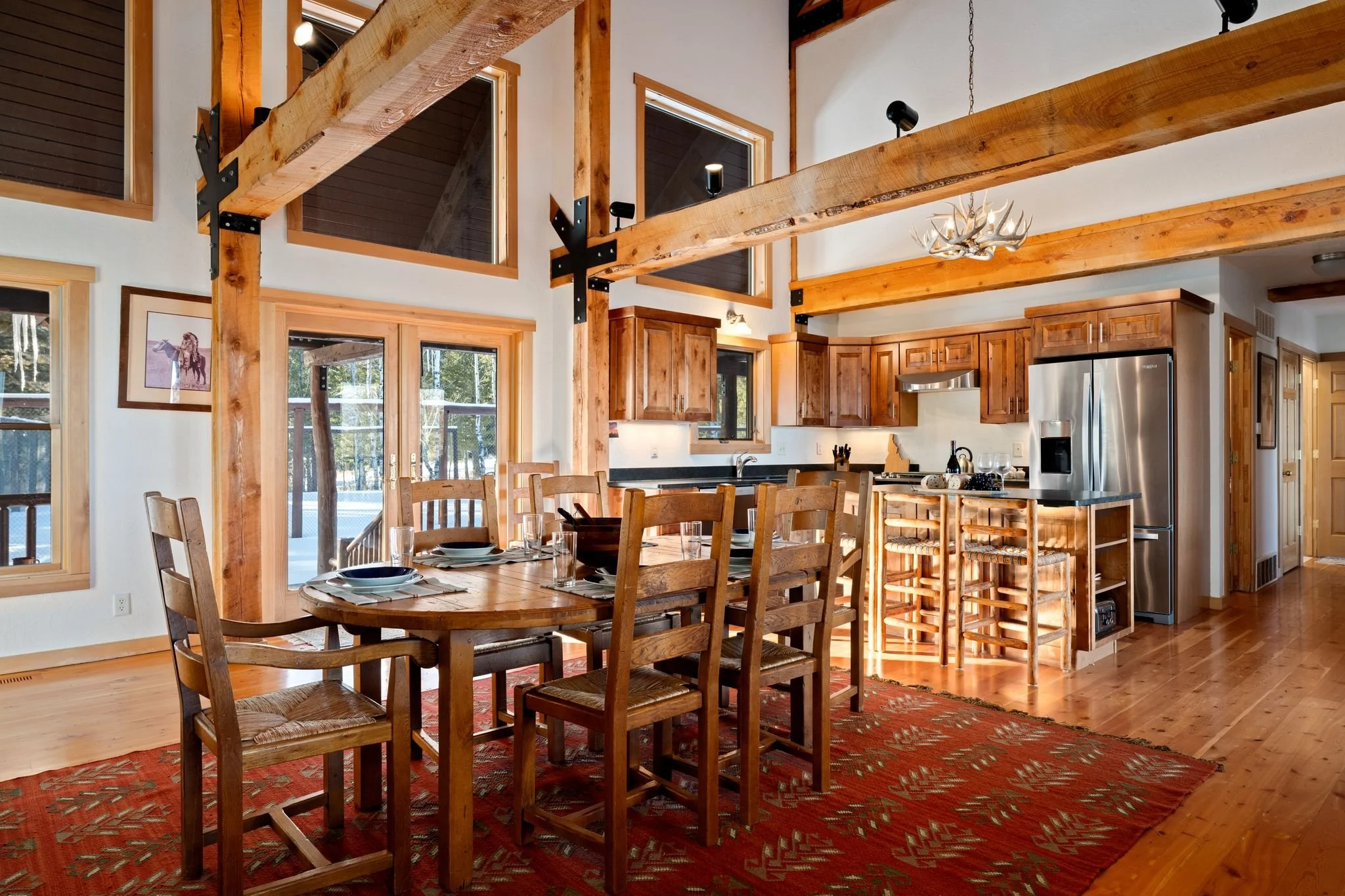 Interior of a rustic kitchen and dining area with wooden beams, a round wooden dining table set with plates and glasses, and a kitchen with wooden cabinets, a stainless steel refrigerator, and bar stools at a kitchen island. Large windows and glass d
