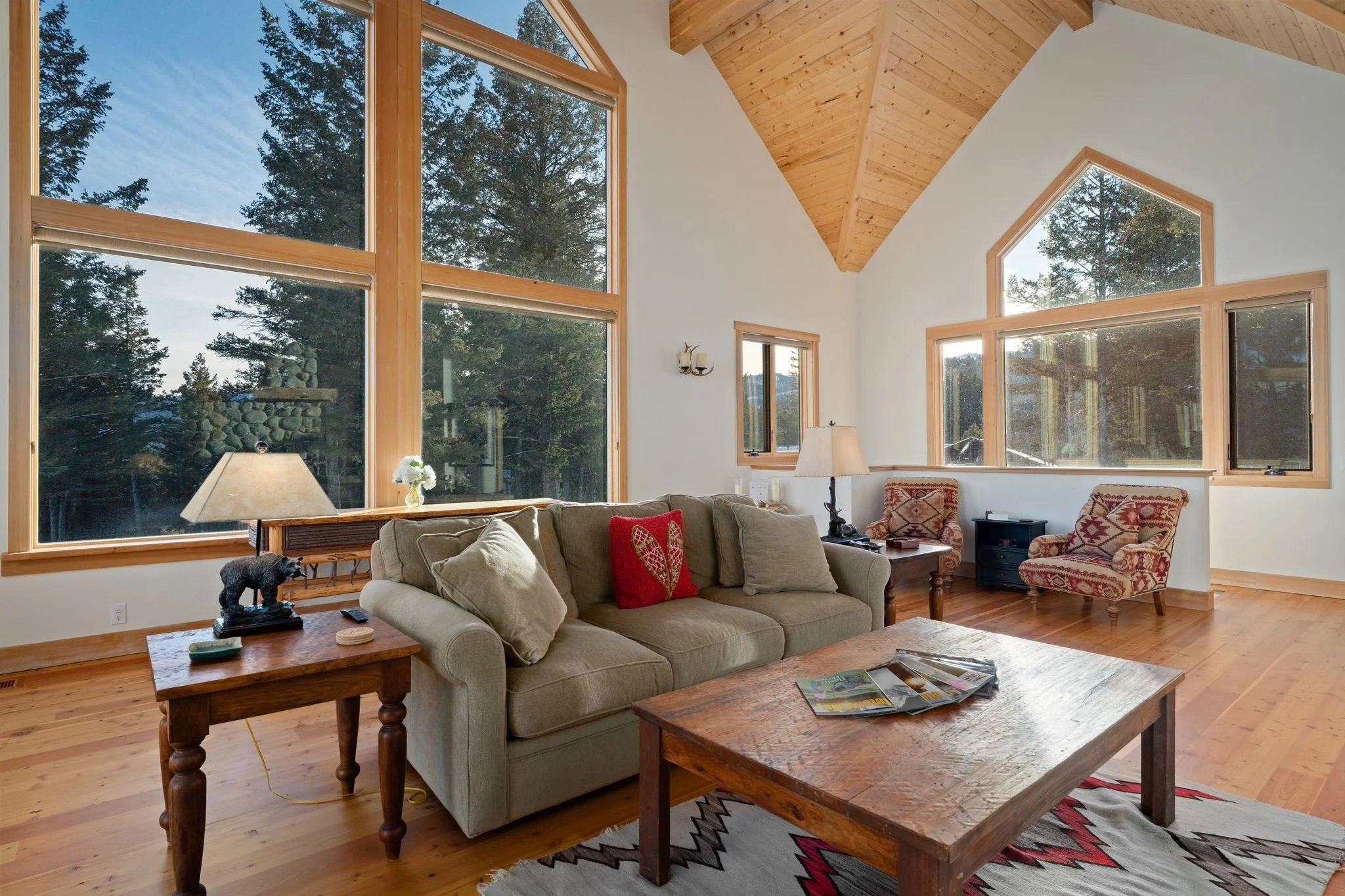 Living room with large windows, wooden ceiling, beige sofa, patterned armchairs, wooden coffee table, side tables, lamps, and decorative items in a mountain cabin style.