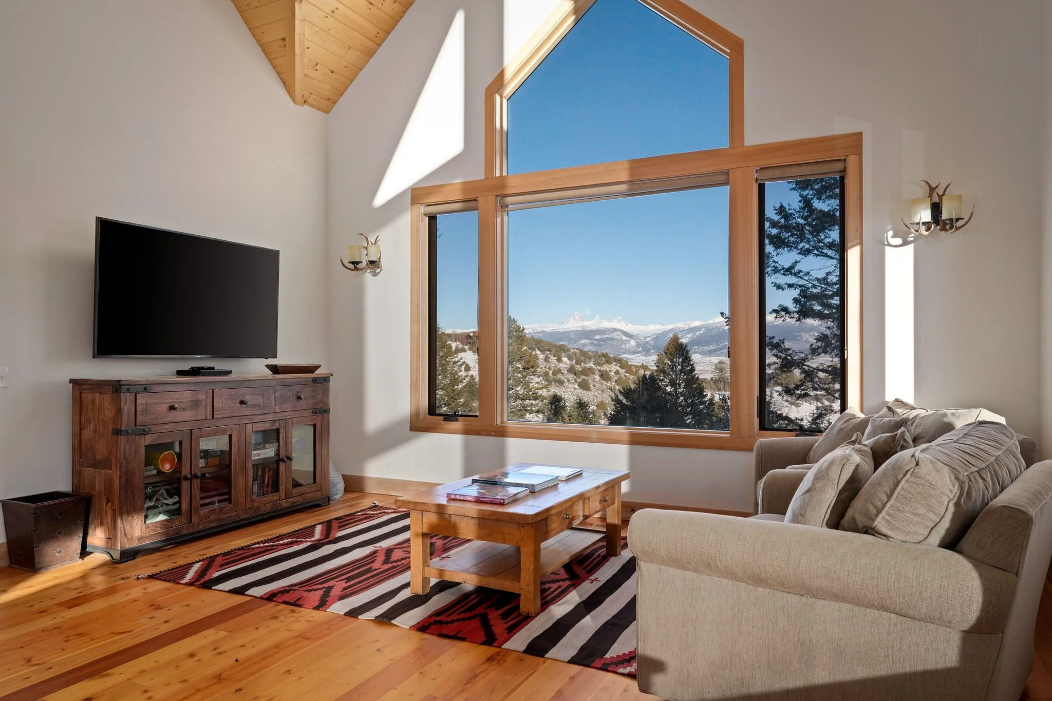 Living room with large window showing snowy mountain view, beige couch, wooden coffee table, TV on wooden stand, colorful rug, wooden ceiling, and wall sconces.