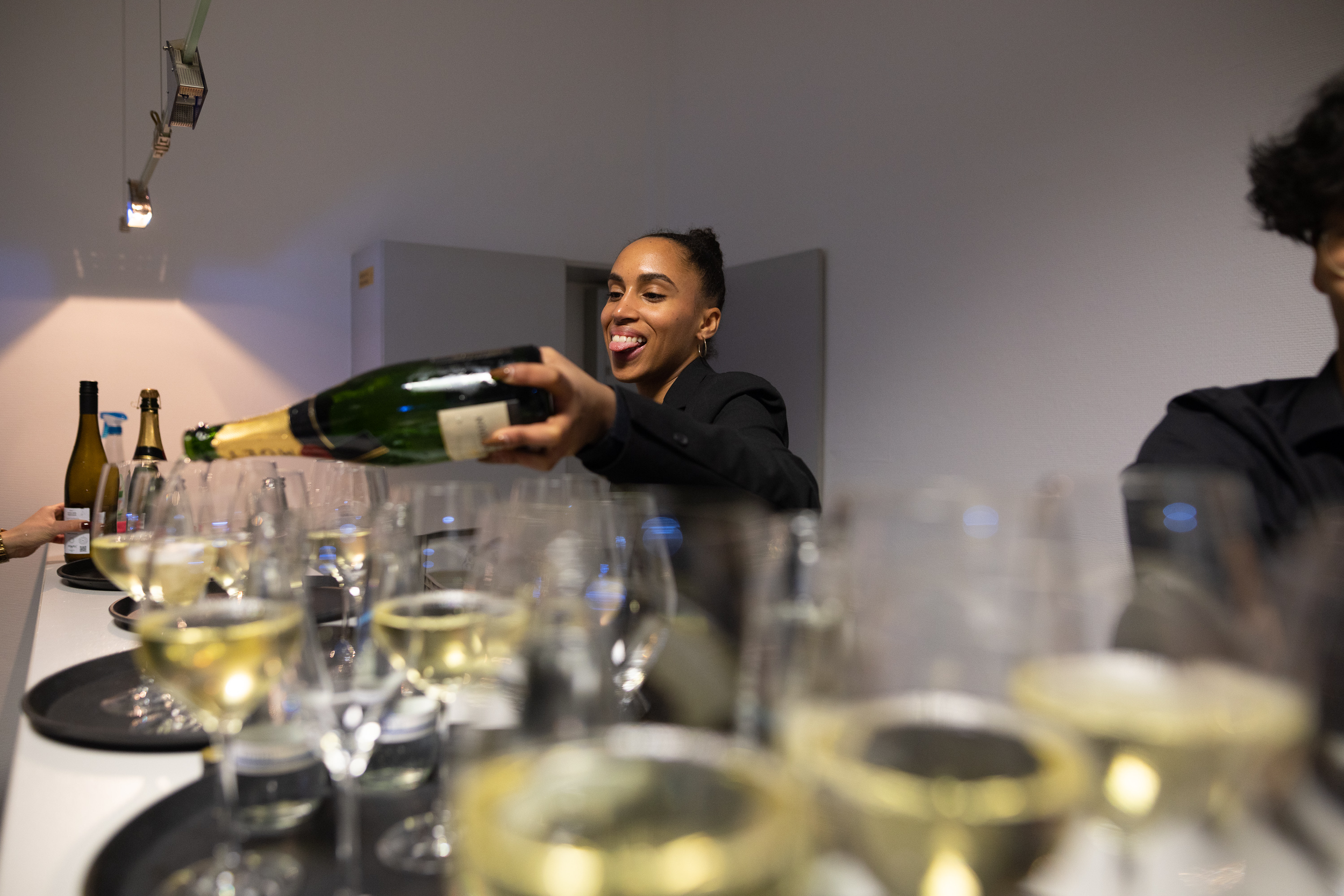 Eine Frau serviert Champagner in Gläsern an einer Veranstaltung. Im Hintergrund sind weitere Flaschen und Gläser auf einem Tisch.