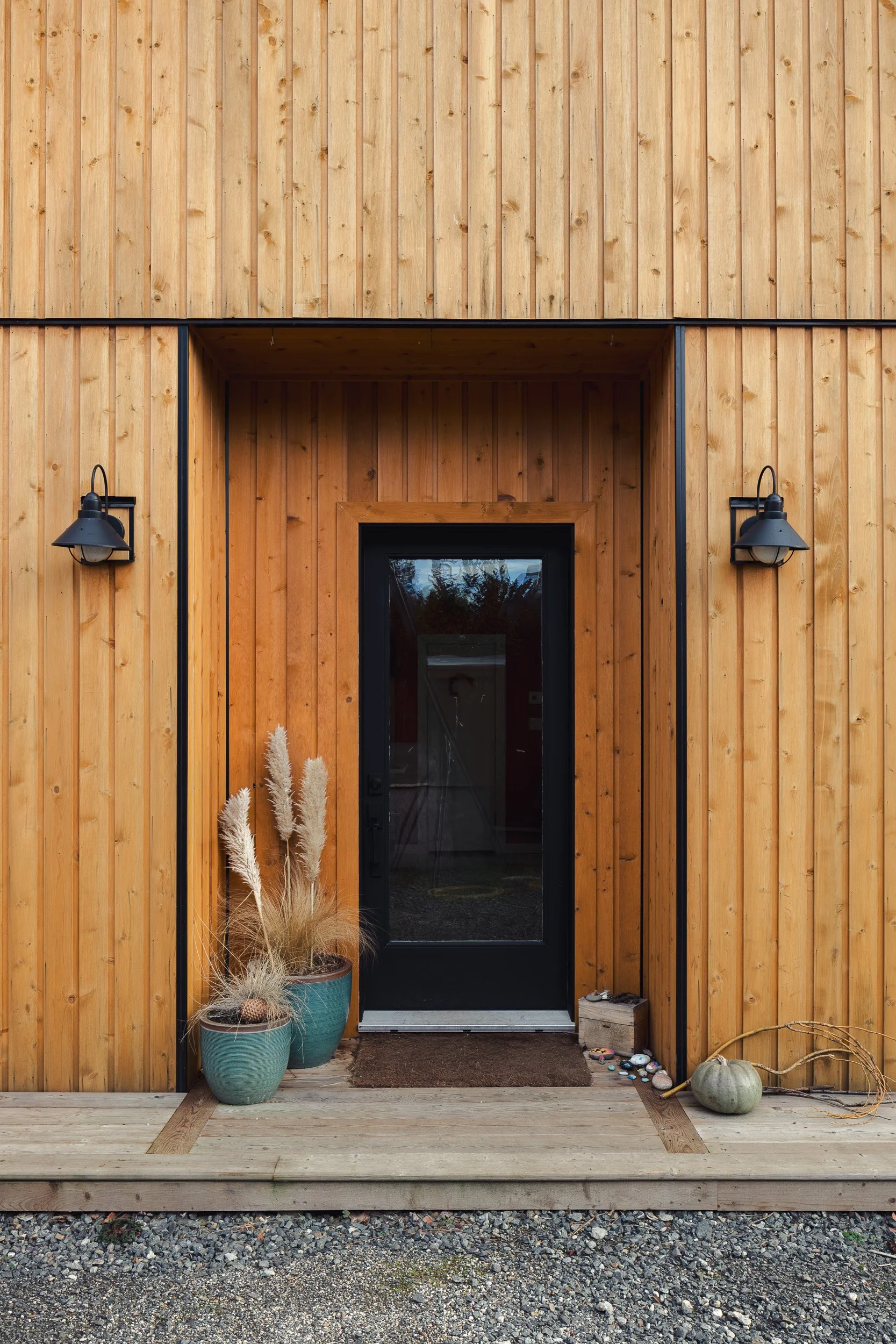 Entrance to a wooden house with a black door, accented by two outdoor wall lamps, potted plants with pampas grass, pumpkins, and small decorative stones.
