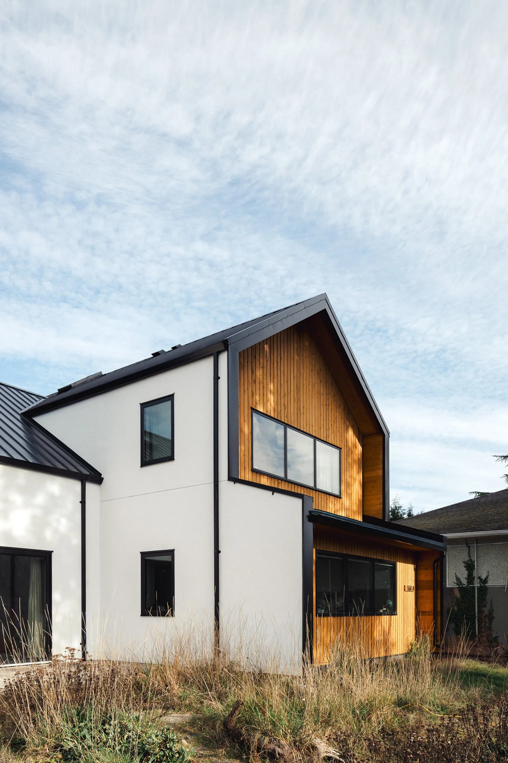 Modern two-story house with white and wooden exterior, black window frames, and a pitched roof, surrounded by tall dry grass under a cloudy sky.