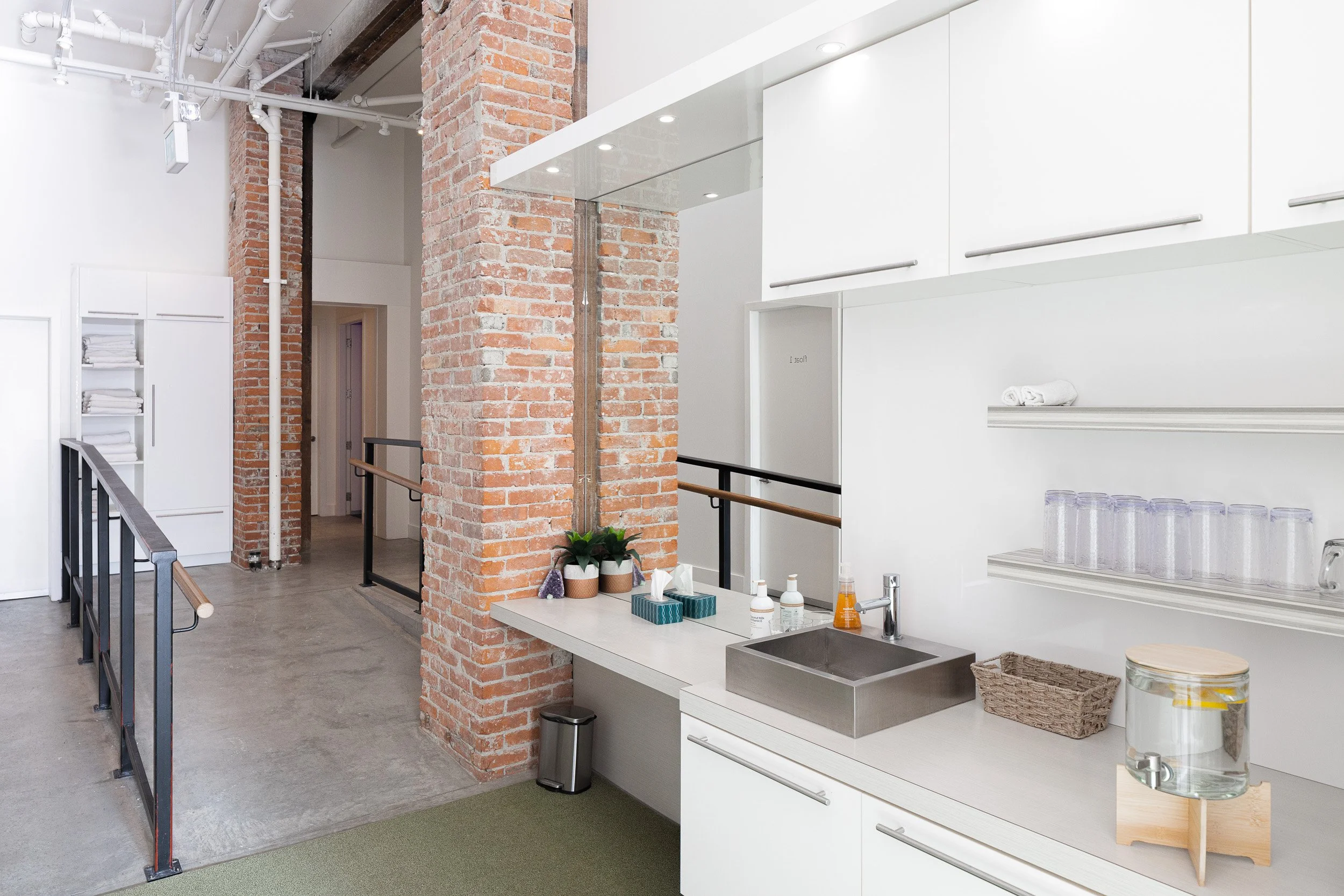 A modern kitchen area with white cabinets, open shelving with glasses, a stainless steel sink, and a countertop with soap dispensers, a water jug, and small plants. The space has exposed brick columns, a concrete floor, and some greenery, with a rail
