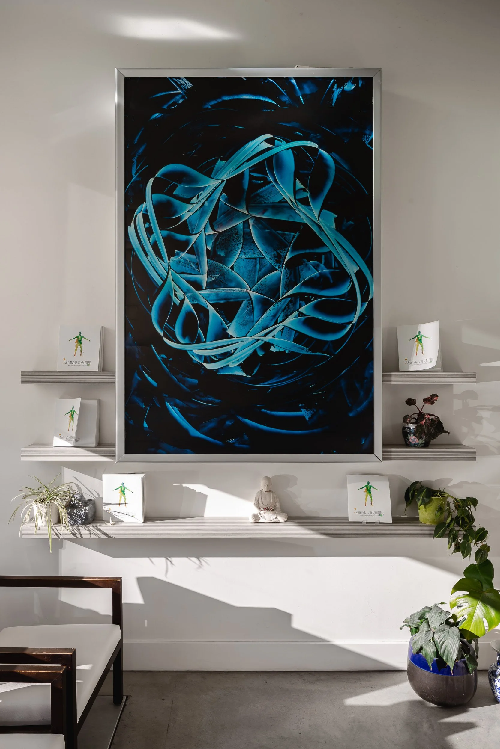 Interior wall with a large abstract digital art piece in shades of blue, surrounded by small potted plants, a small statue, and framed cards with a human figure on white shelves.