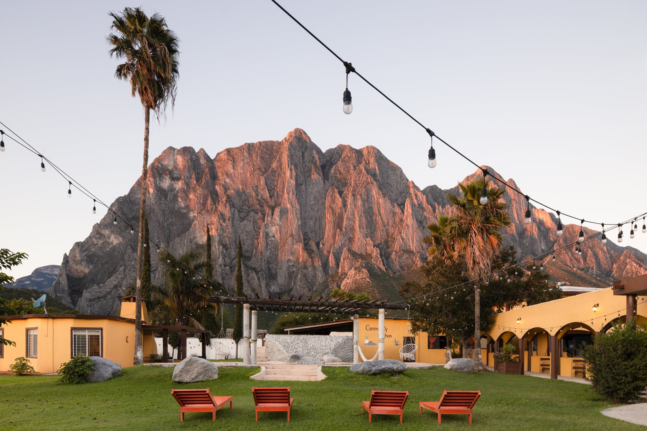 Outdoor scene with green lawn, four orange lounge chairs, large rocks, palm trees, buildings with yellow walls, string lights, and a mountain range in the background during sunset.