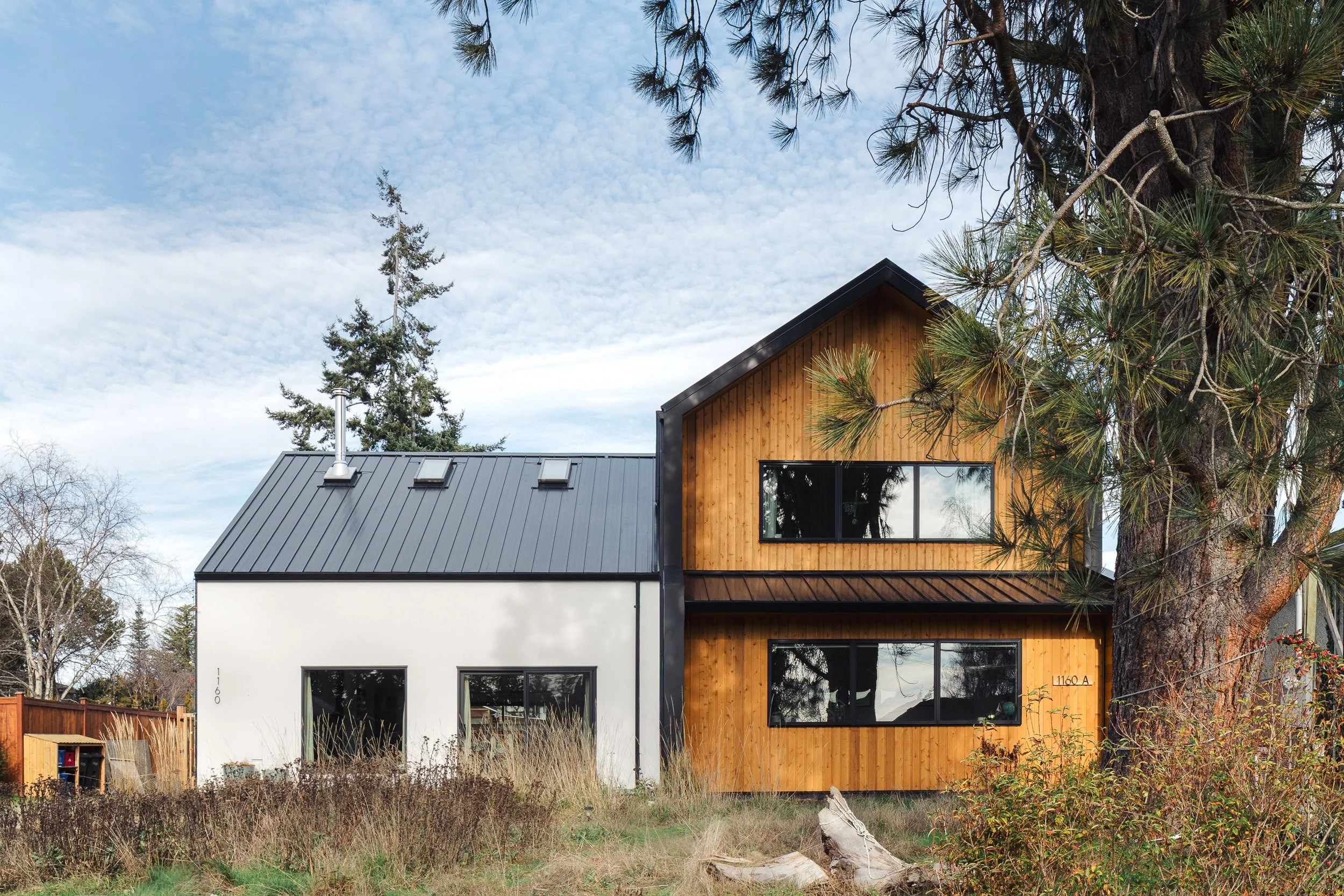 A modern two-story house with a combination of white and natural wood exterior, large black-framed windows, and a dark metal roof, set amidst trees and tall grass.