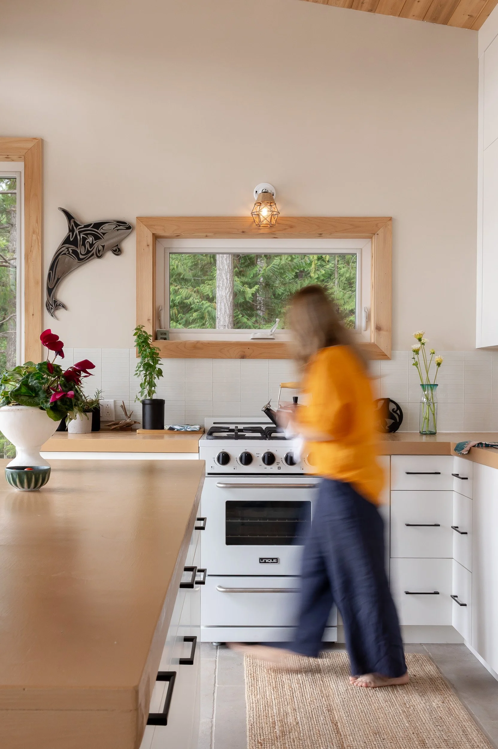 A woman in yellow top and dark pants walking barefoot in a modern kitchen with white cabinets, wooden countertops, a stove, and a window showing trees outside.