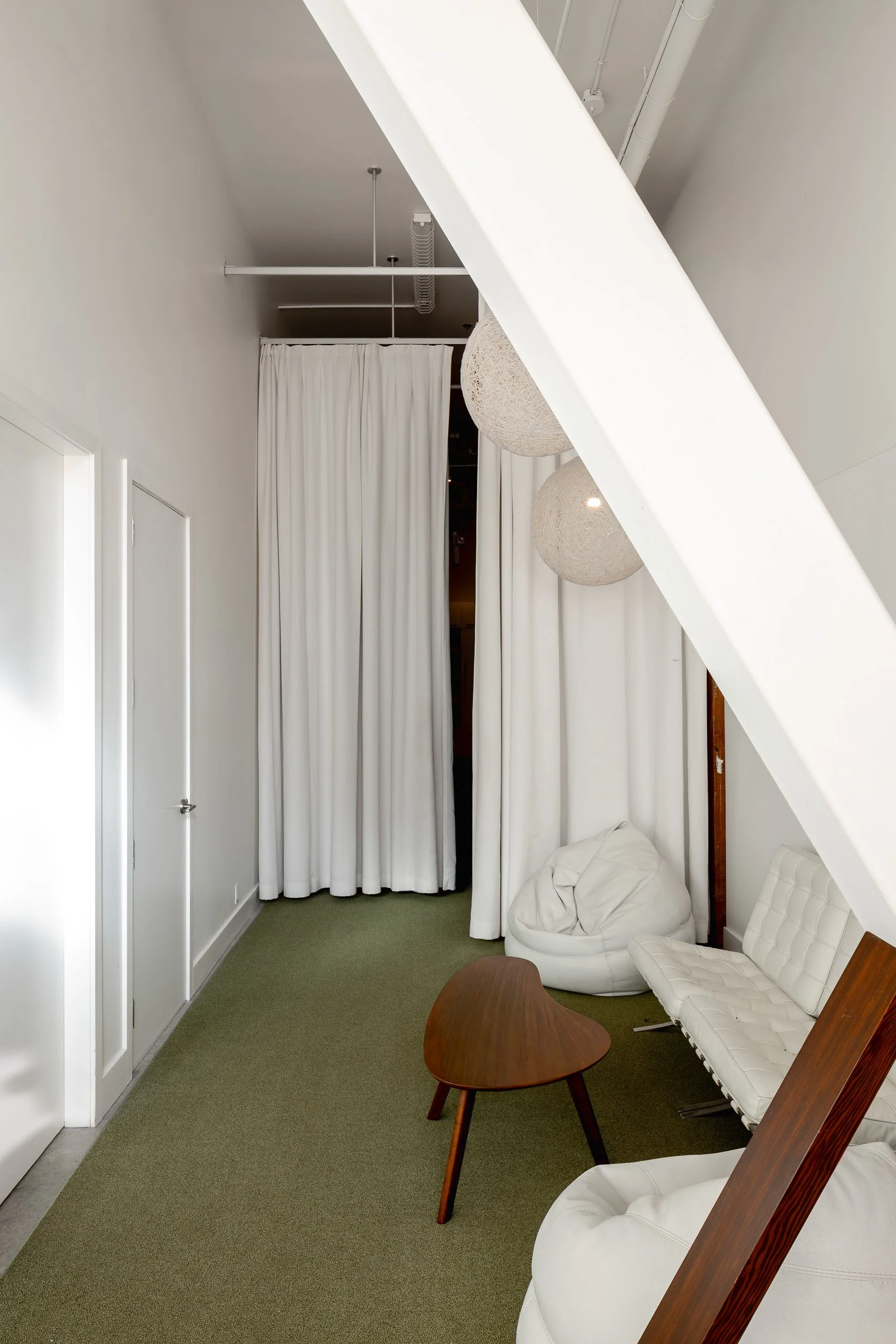 Modern minimalist interior with white curtains, green carpet, wooden coffee table, white leather chair, and hanging spherical light fixtures.