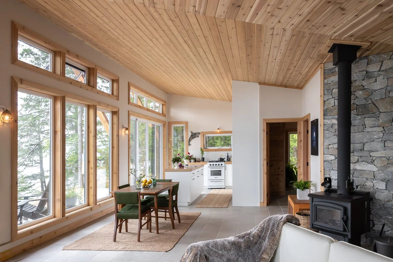 Interior view of a cozy, rustic-style living space with large windows, a dining table with green chairs, a kitchen with white cabinets, and a stone fireplace with a black stove