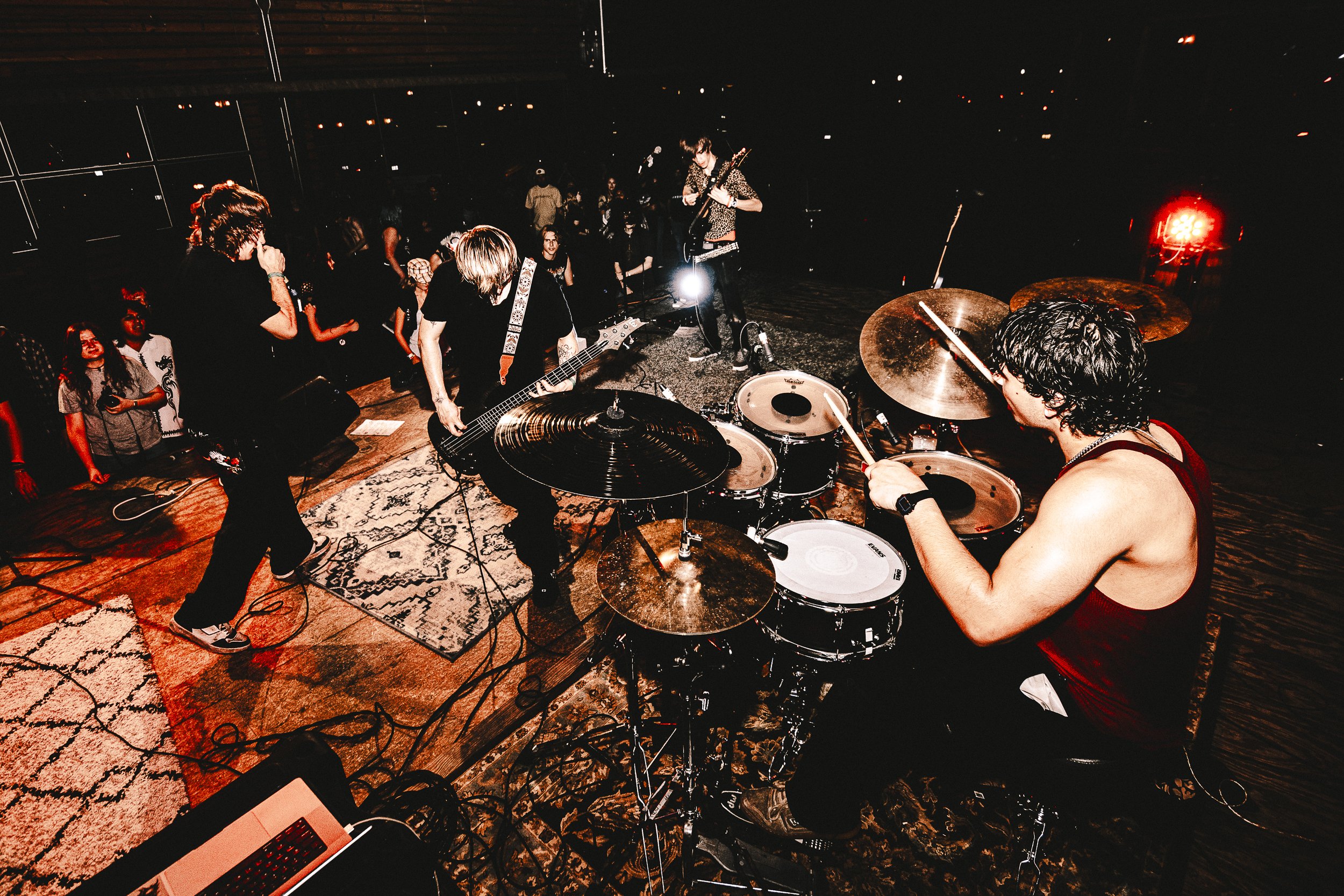A live band performing on stage with a drummer, guitarist, and vocalist, while the audience watches in a dimly lit venue with stage lights.