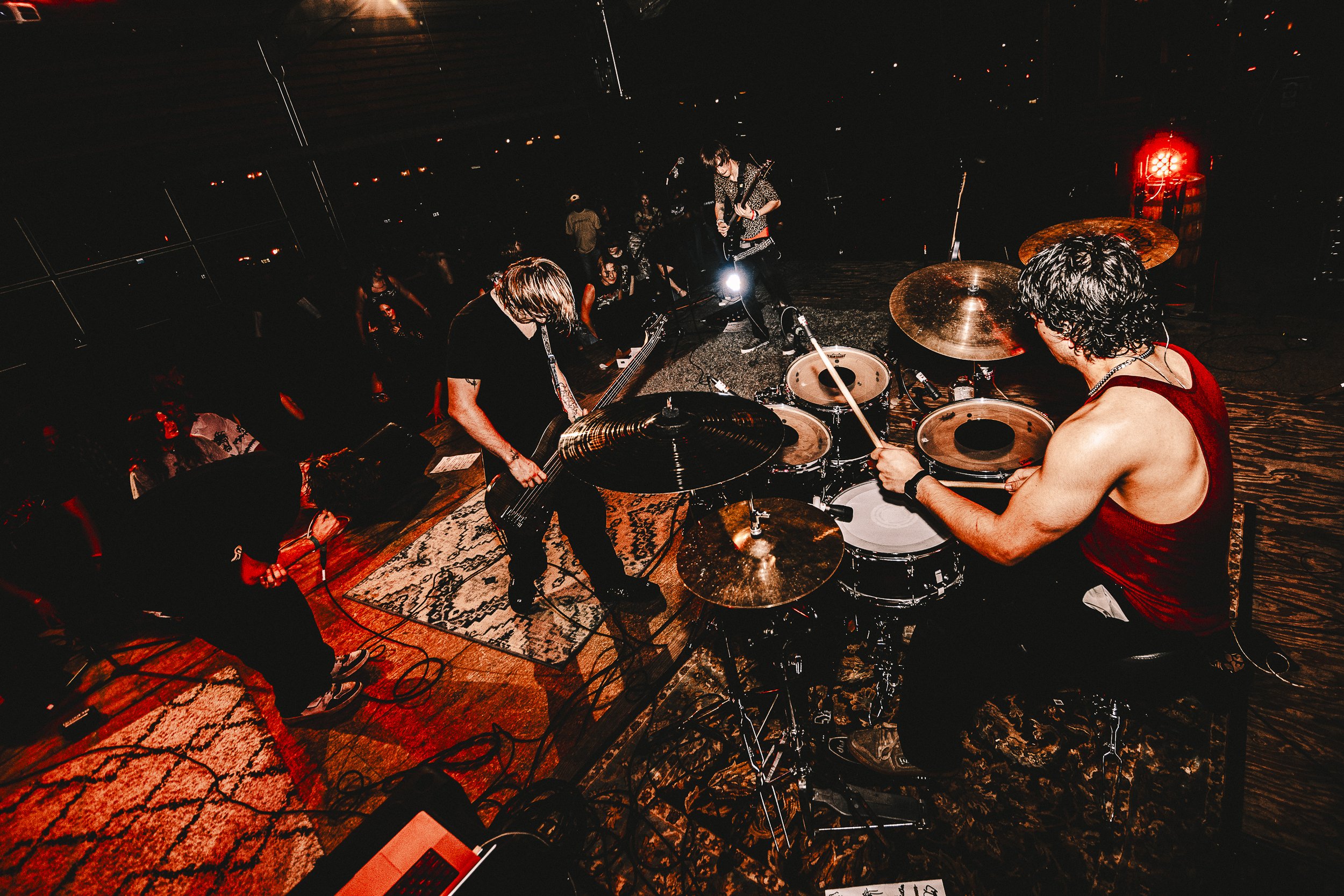 View of a band performing on stage, including a drummer, guitarist, and other musicians, with audience in the background.