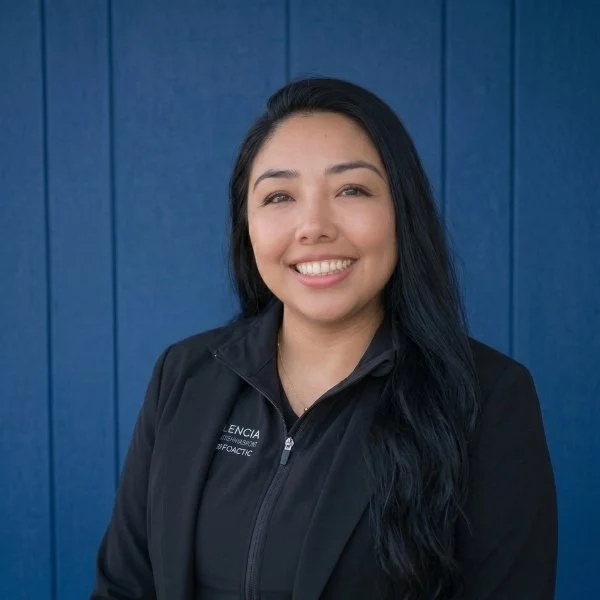 Young woman with long dark hair smiling, wearing a black jacket, standing in front of a blue wall.
