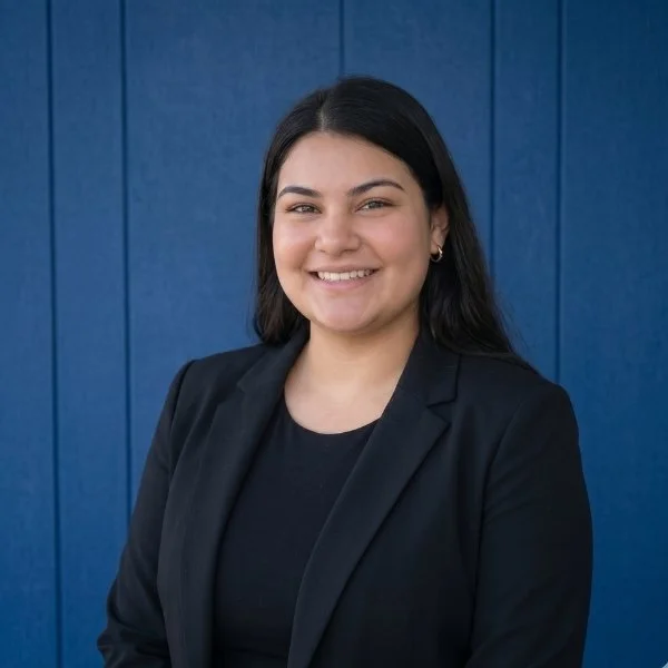 A young woman with dark hair, wearing a black blazer and black shirt, smiling in front of a blue wall.