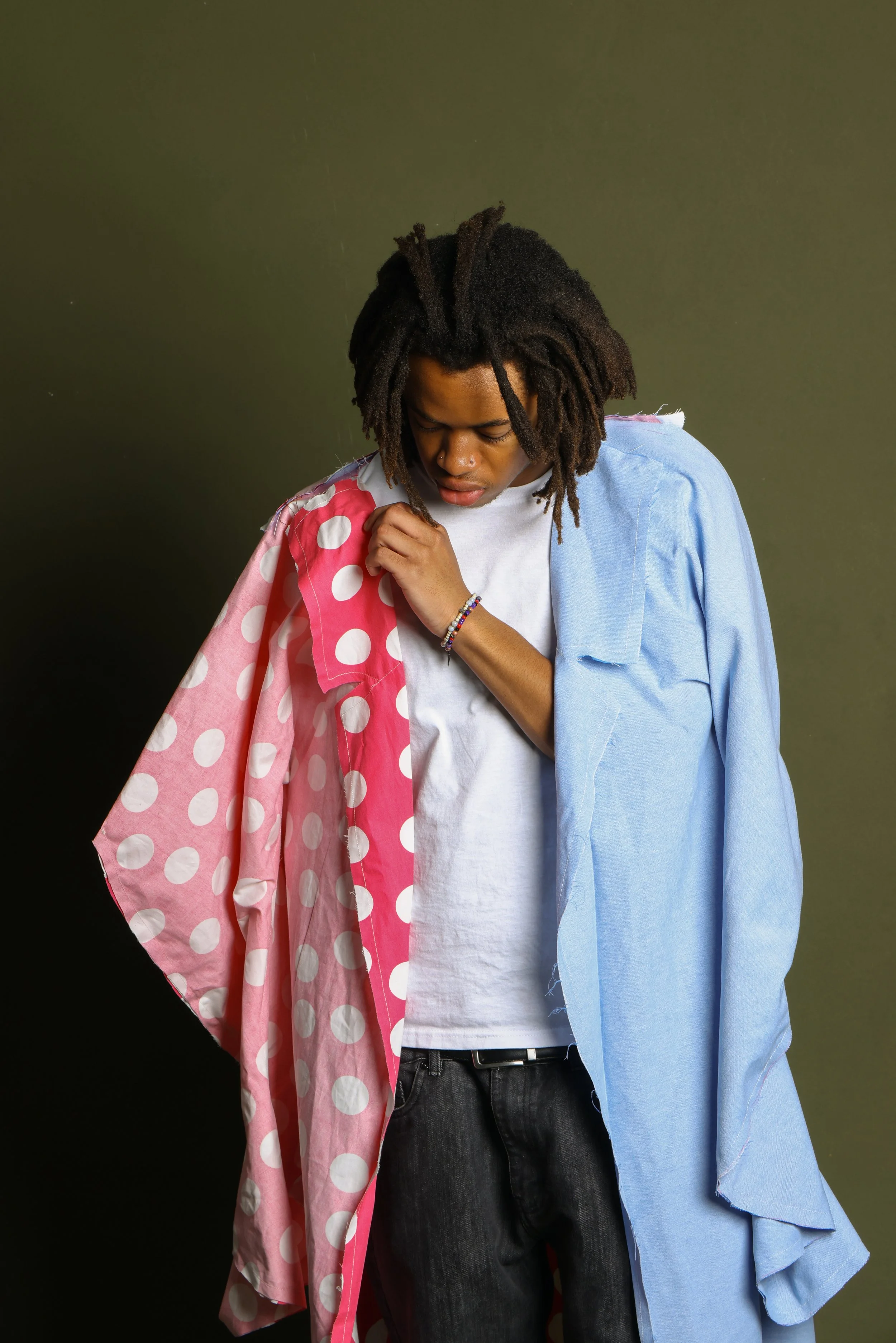 A young man with dreadlocks is wearing a white t-shirt, black jeans, and a light blue jacket draped over his shoulder. He is holding a pink scarf with white polka dots against the jacket, looking down with a contemplative expression.