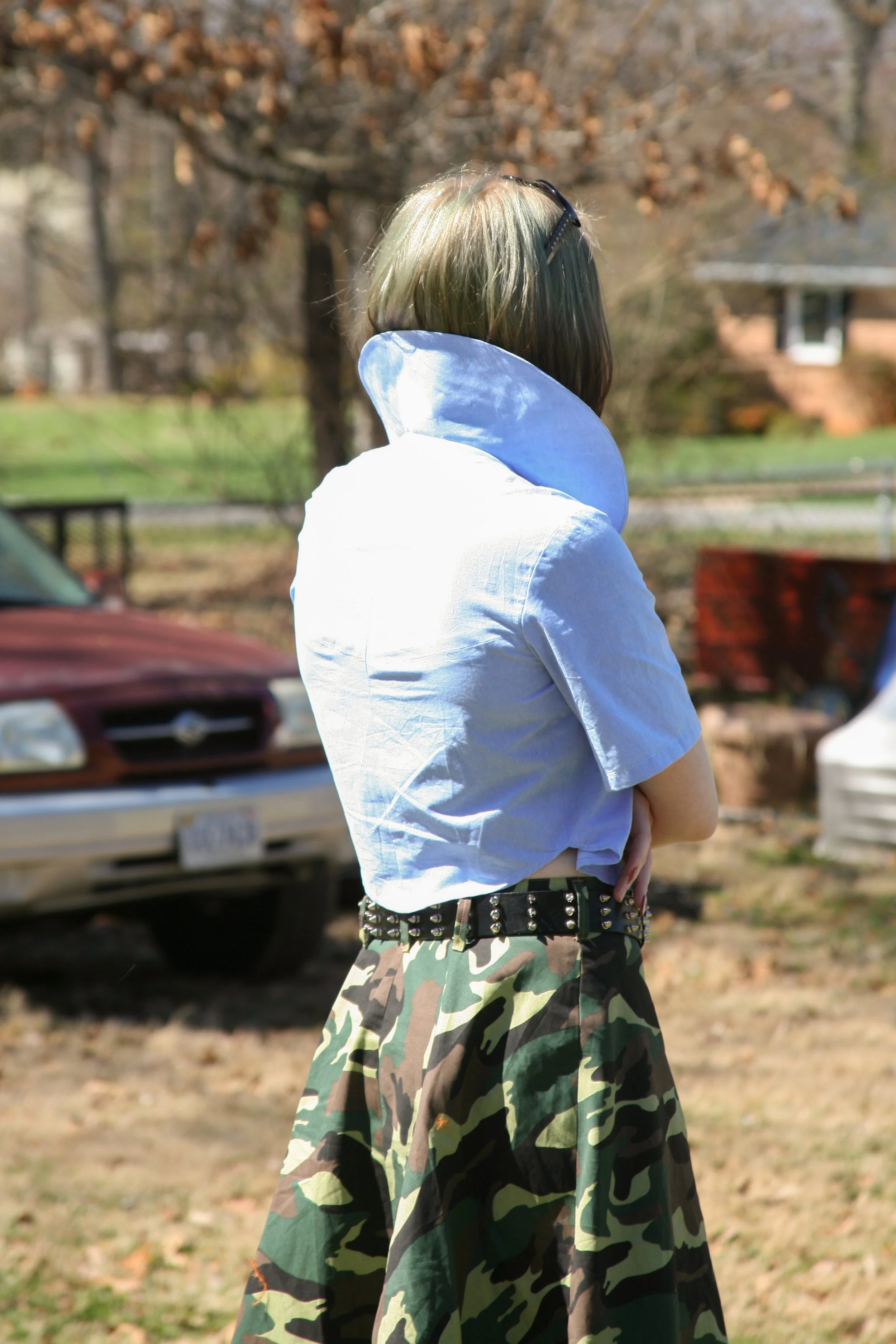Person with green hair wearing sunglasses, a white shirt with a high collar, and a camouflage skirt standing outdoors with arms crossed, with an old red car and trees in the background.