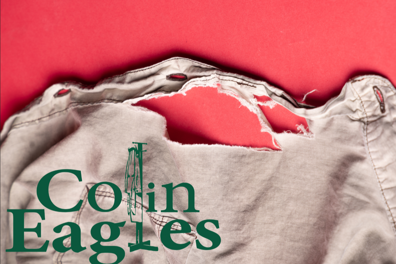 Close-up of a torn pair of khaki shorts with an American flag and the words "Coin Eagles" printed on the pocket area.