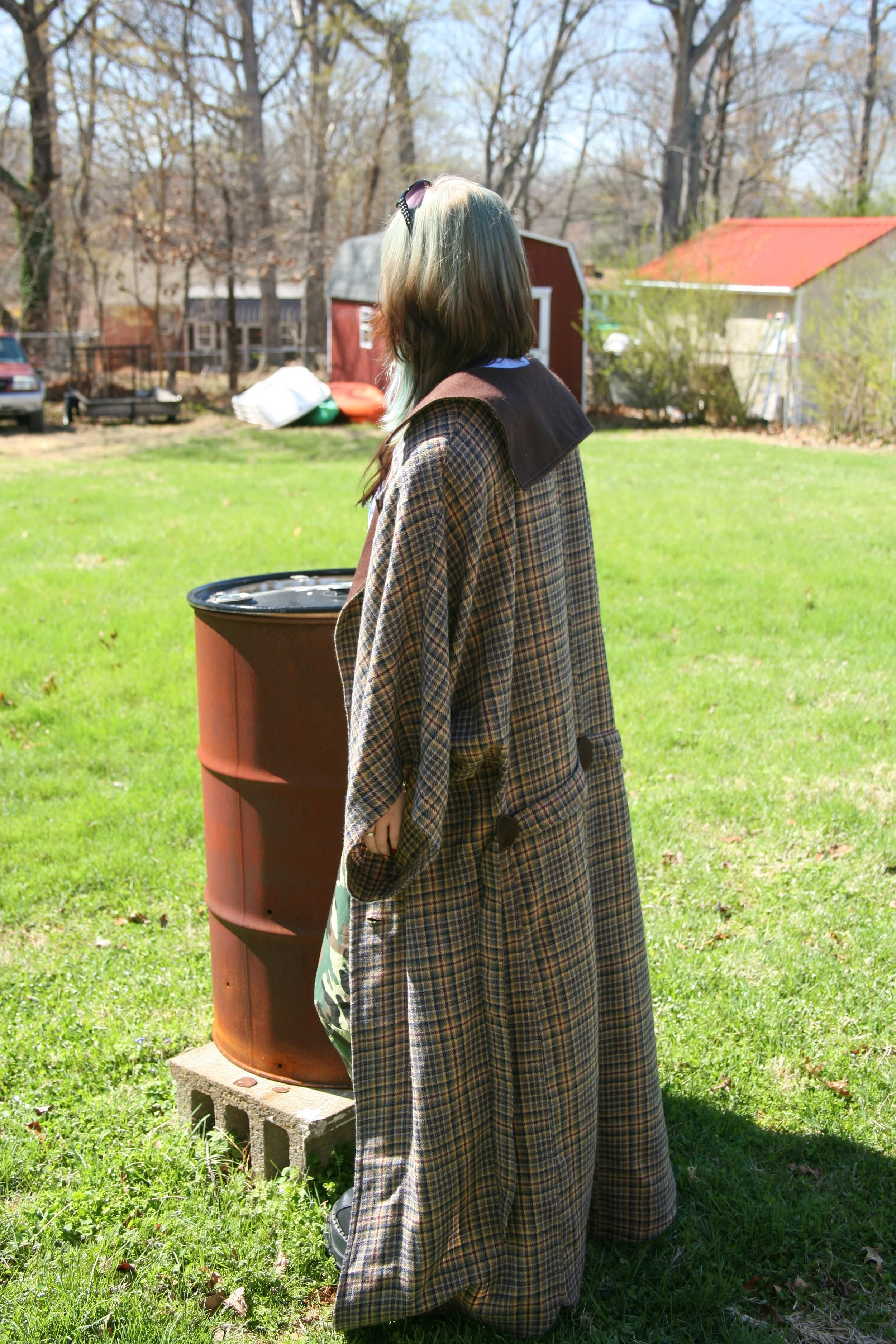 A person with long hair wearing a brown plaid coat standing outdoors in a yard, looking at a rusty barrel.