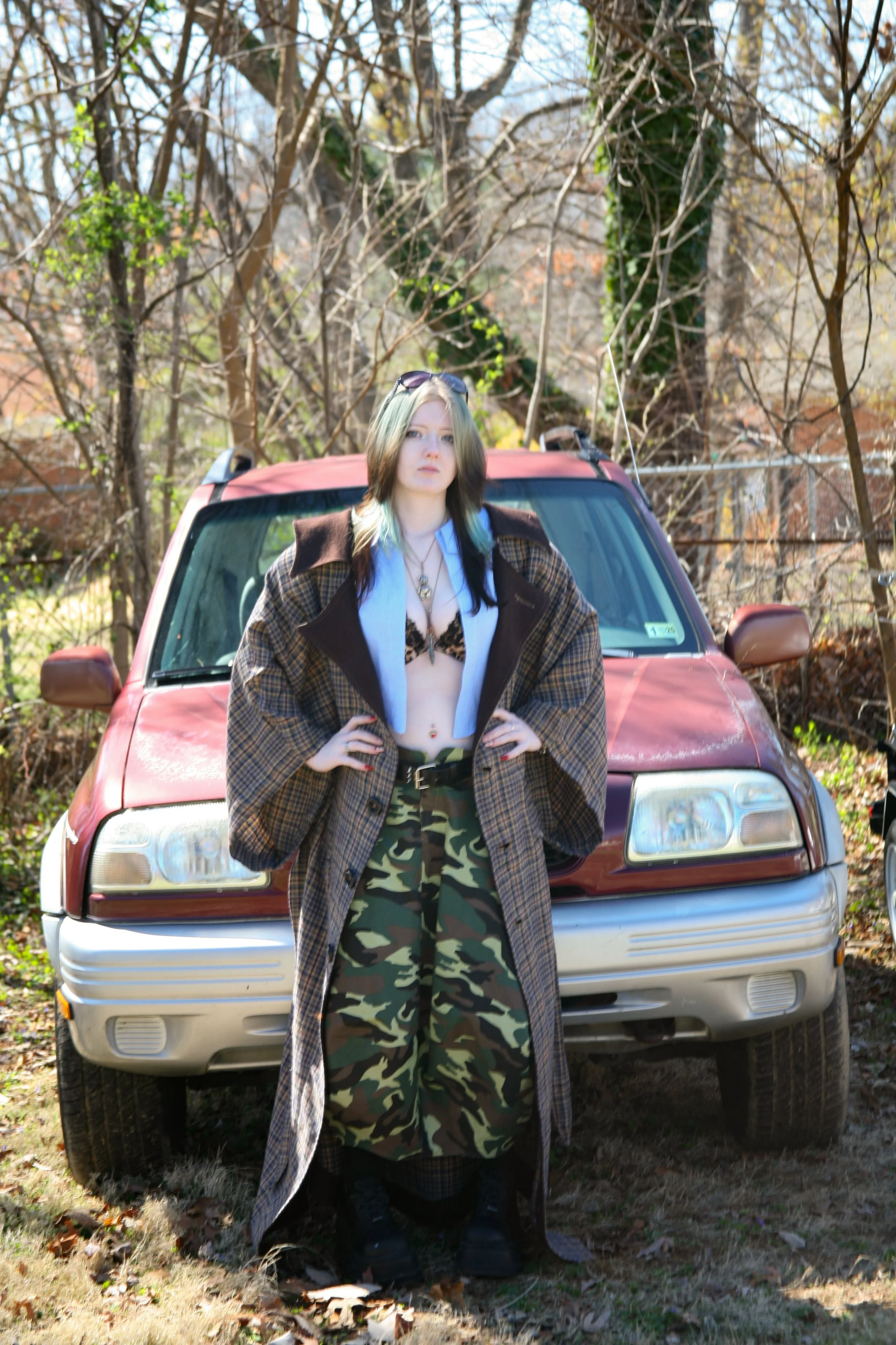 A young woman standing in front of an older red SUV car in a wooded area with leafless trees.