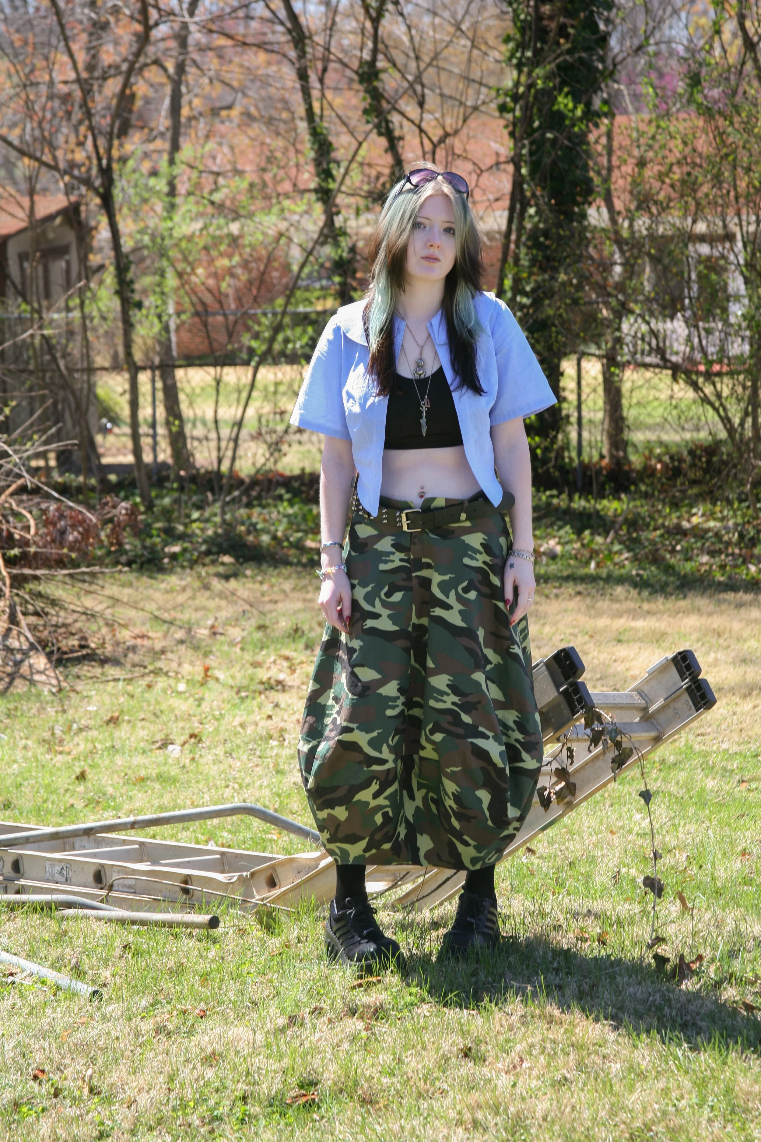 Young woman standing outdoors on grass in a backyard, wearing a light blue short-sleeve shirt, black crop top, camouflage baggy pants, black sneakers, and sunglasses on her head, with trees and houses in the background.