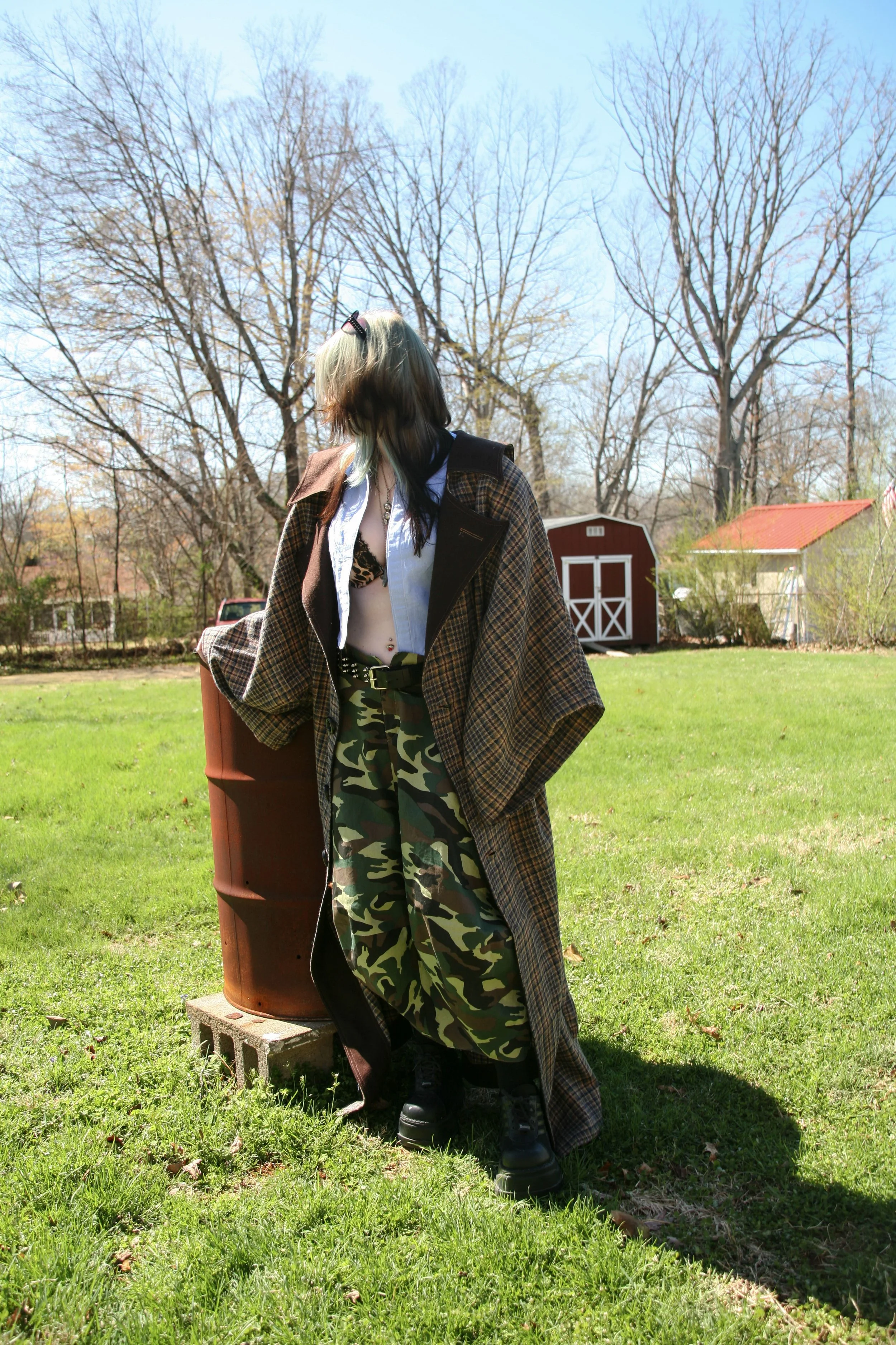 Person standing outdoors on a grassy lawn, dressed in camouflage pants, black boots, and a long plaid coat, with trees and a red barn in the background.