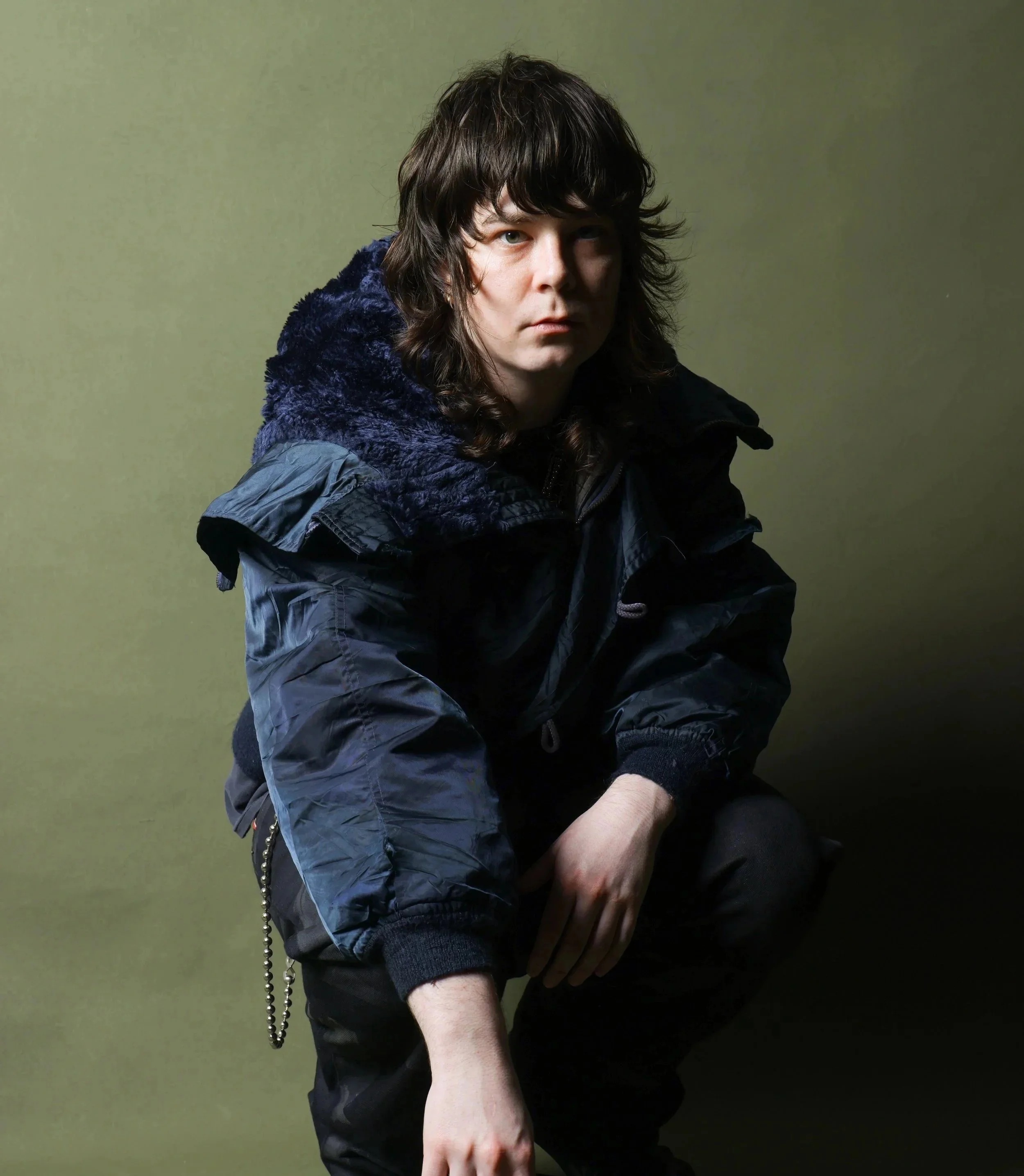 A young man with long dark hair and fair skin squatting against a plain olive-green background, wearing a dark hooded jacket and black pants.