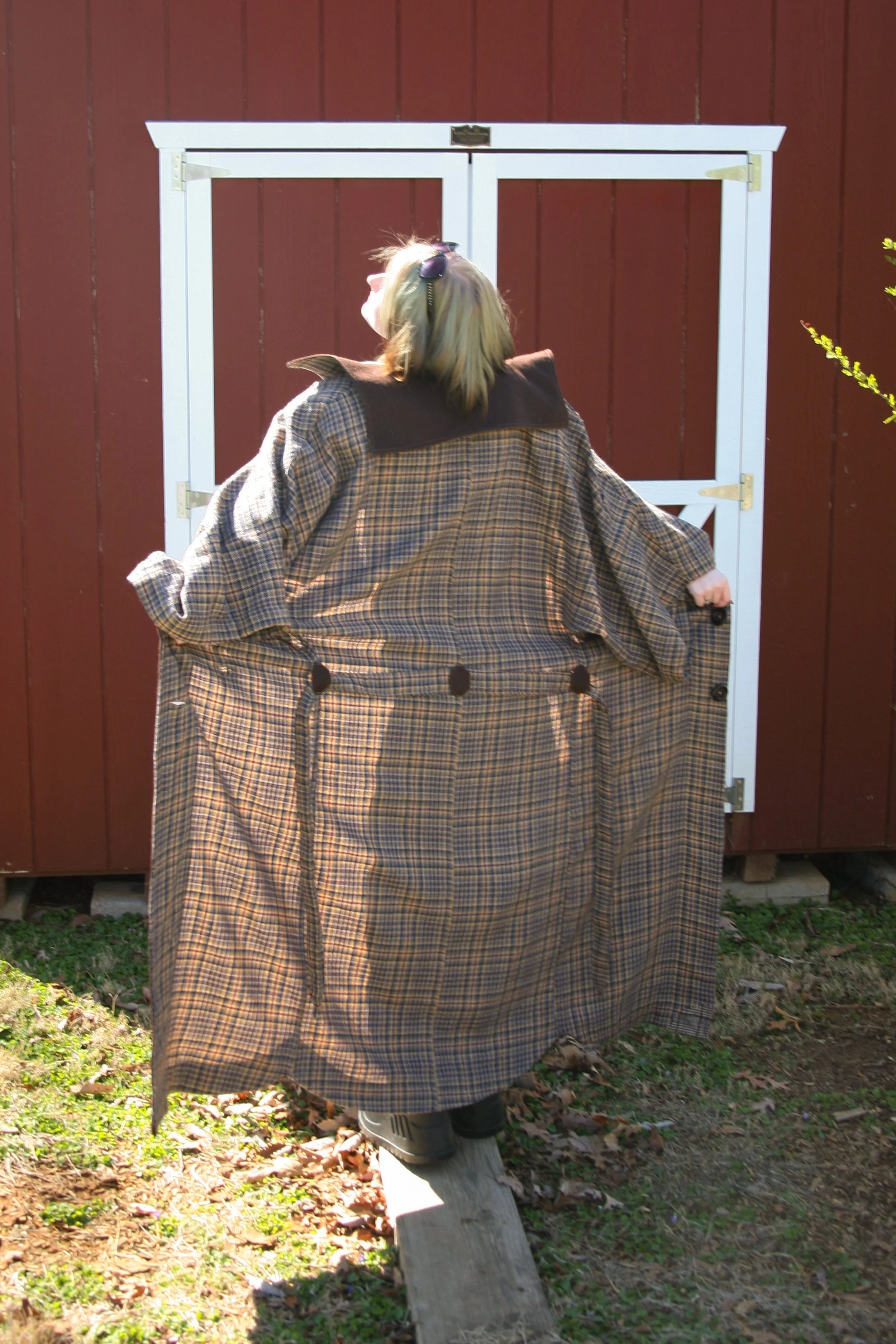 A person with blonde hair wearing a plaid coat and black boots standing outside in front of a red wooden wall, reaching into a large plaid coat or blanket.