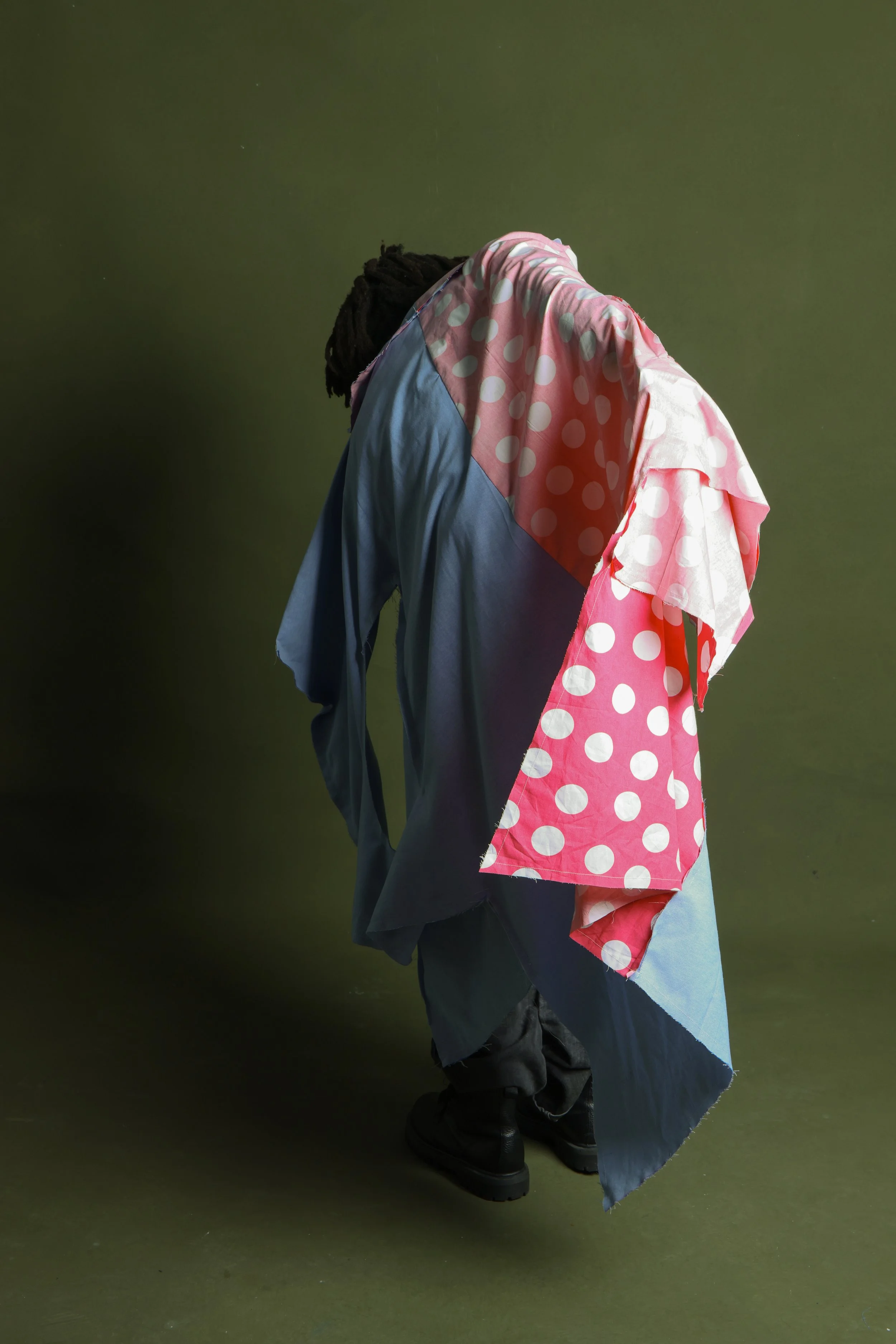 Person wearing layered, colorful, patterned clothing with polka dots and solid colors, standing against a plain green background.