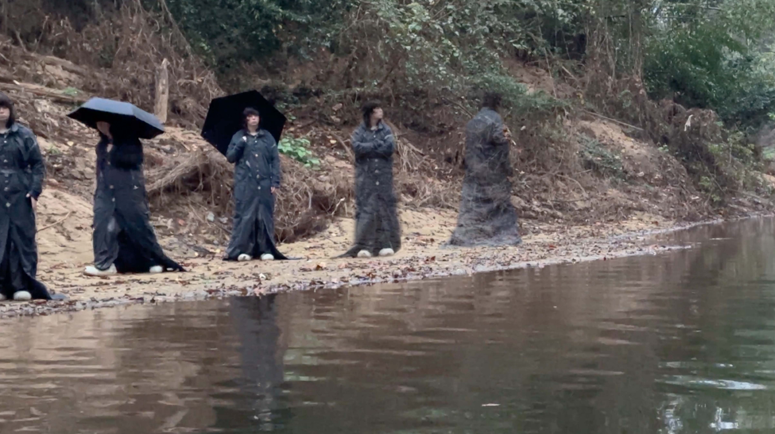 A group of five people wearing raincoats and holding umbrellas standing on a sandy beach near the water's edge. The shoreline is lined with trees and bushes.