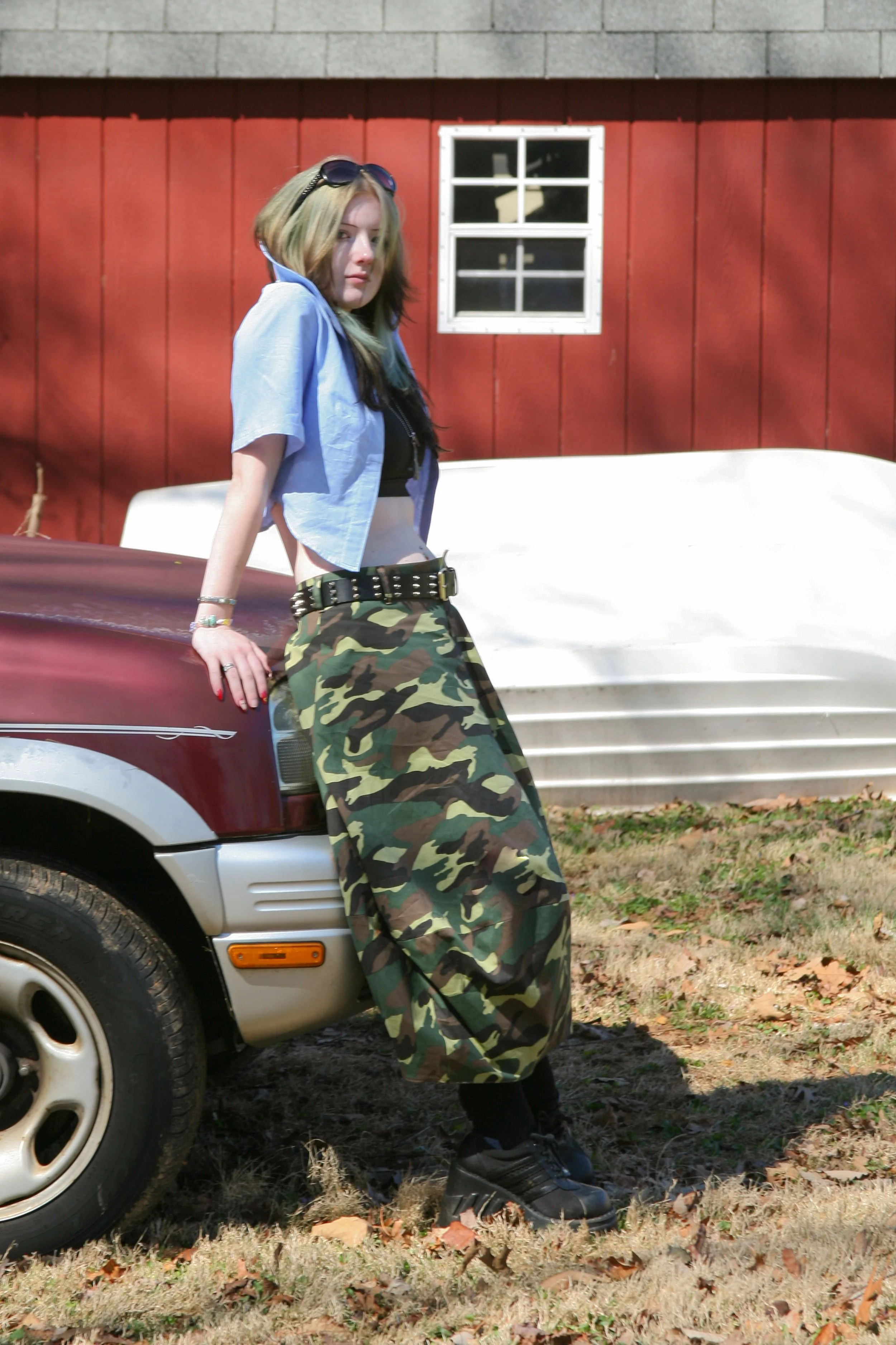 A young woman leaning against a car, wearing camo pants, a light blue cropped jacket, black shoes, and sunglasses on her head, in front of a red barn with a small window.