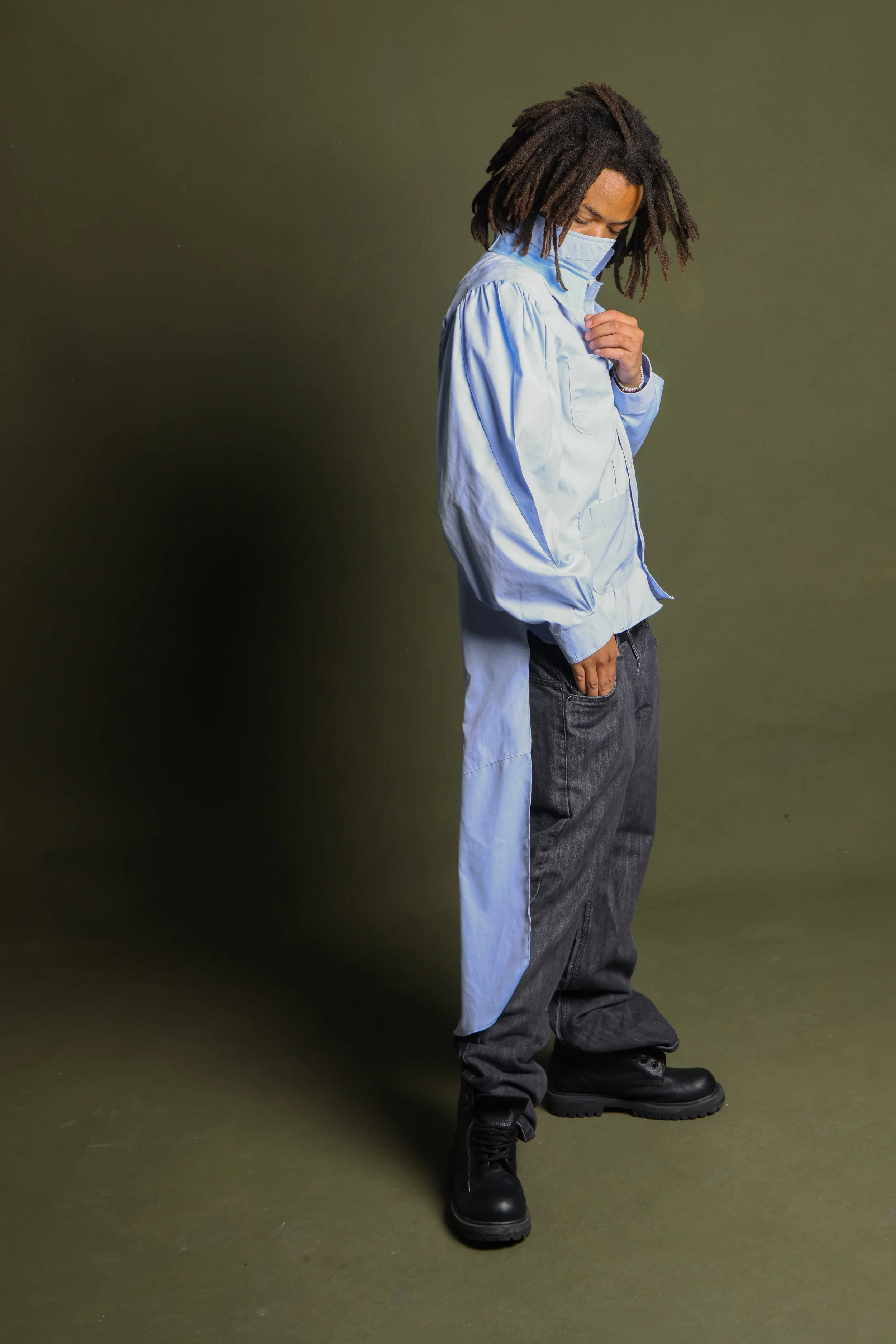 A person with dreadlocks wearing a light blue jacket, dark gray pants, black boots, and a face mask, standing against an olive green background.