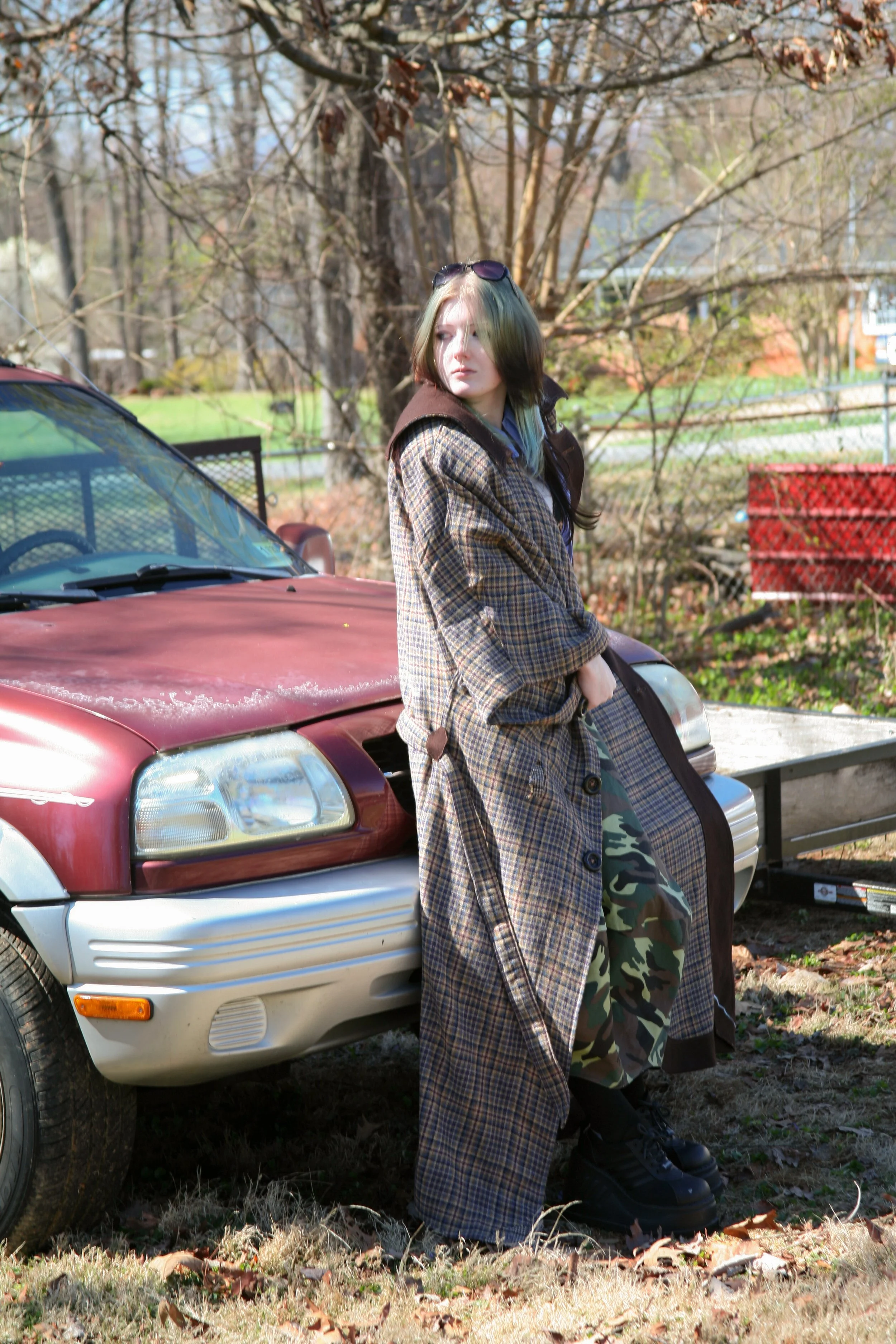 Young woman leaning against a red car in a yard in spring, wearing a large plaid coat, camouflage pants, and black boots, with sunglasses on her head and trees in the background.
