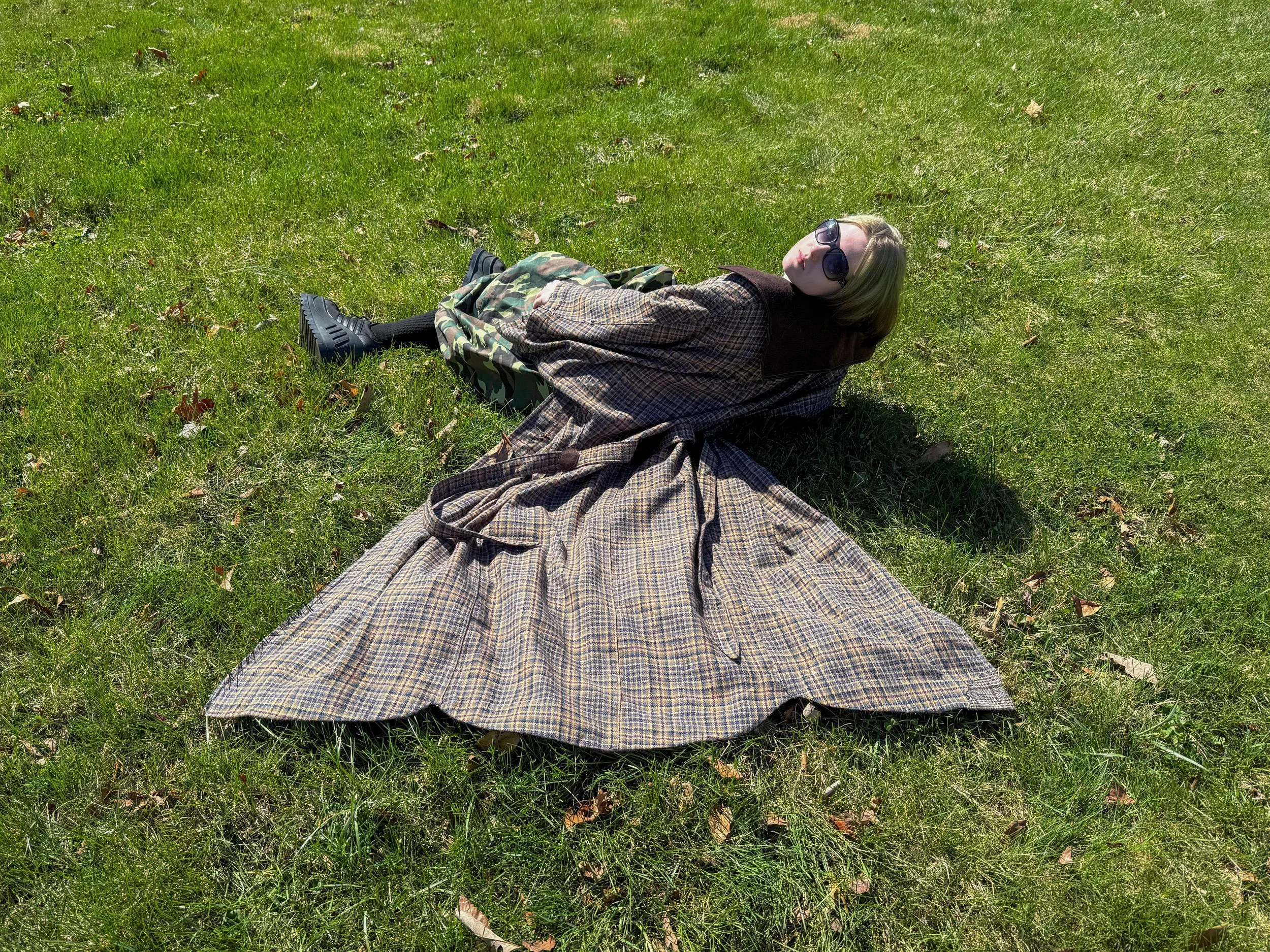 Person lying on green grass, wearing sunglasses, a plaid coat, camo pants, and black shoes, basking in sunlight.