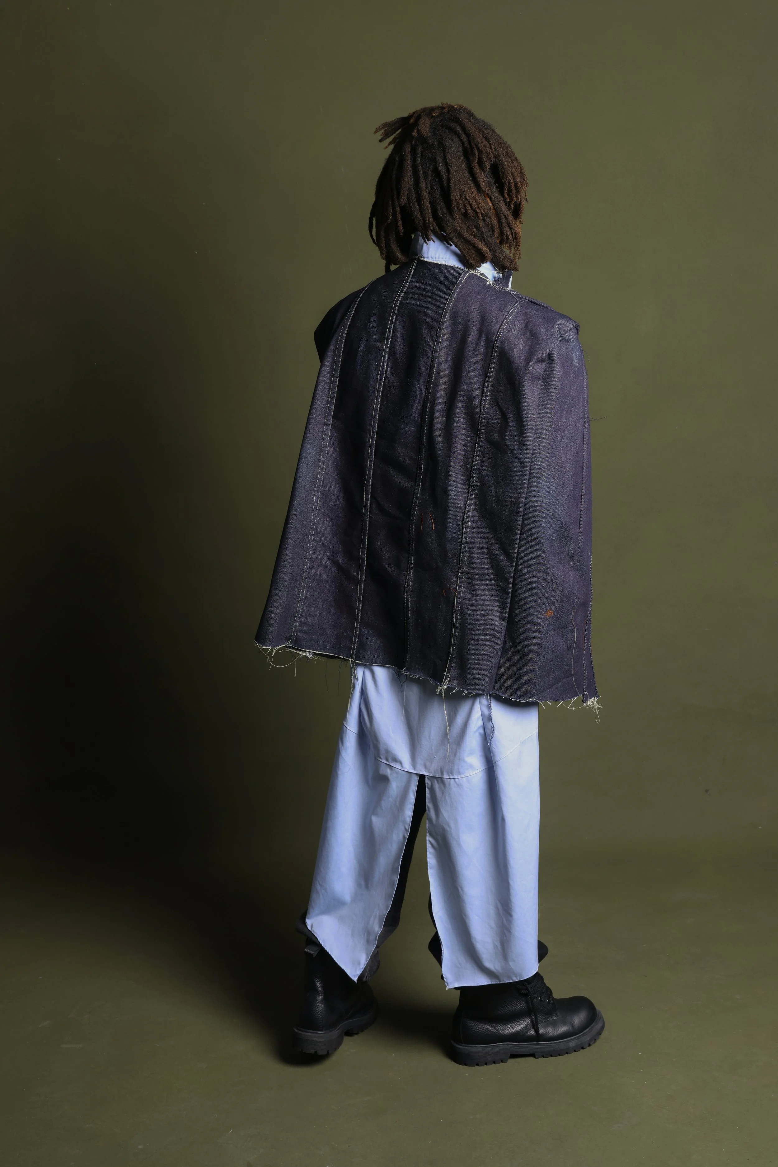 Person standing with their back to the camera, wearing a dark oversized jacket, light blue wide-leg pants, and black boots, against a plain olive-green background.